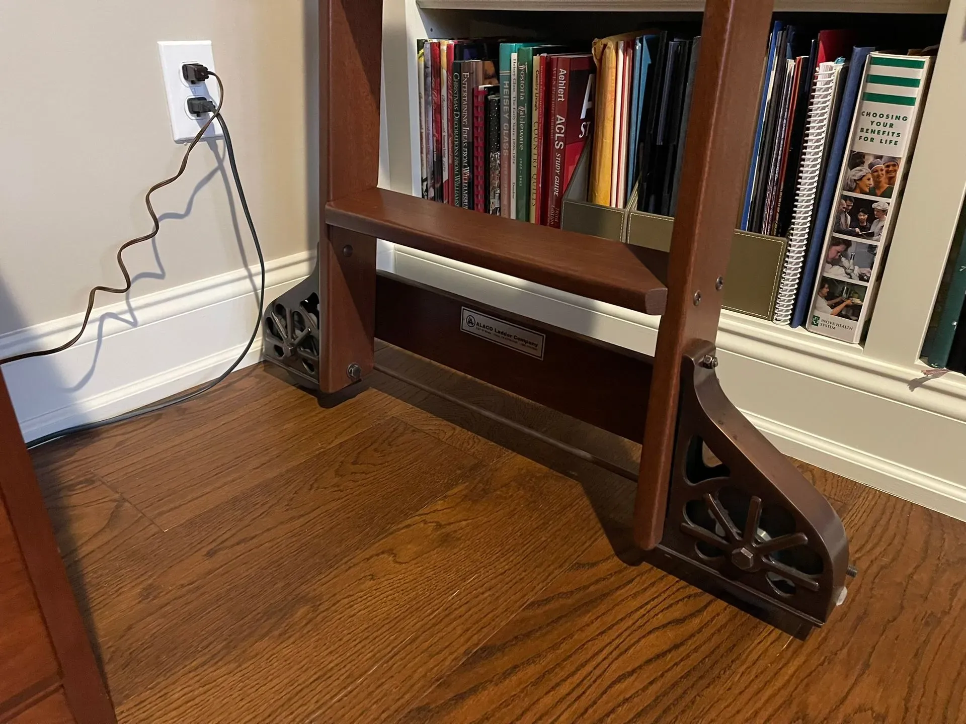 Wooden bookcase with books beside a wall outlet and baseboard in a carpeted room