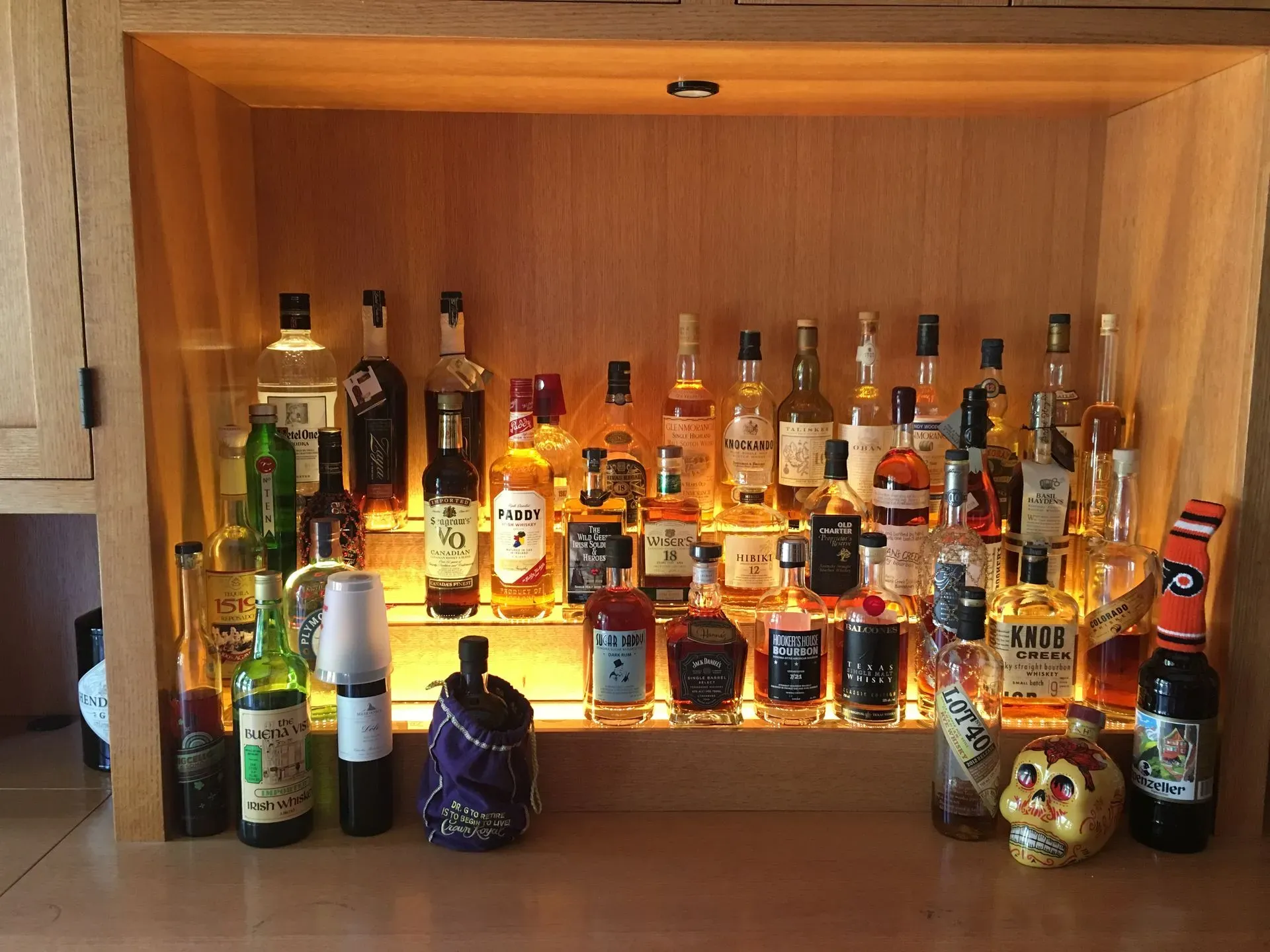 Backlit shelf of assorted liquor bottles and glassware in a wooden cabinet