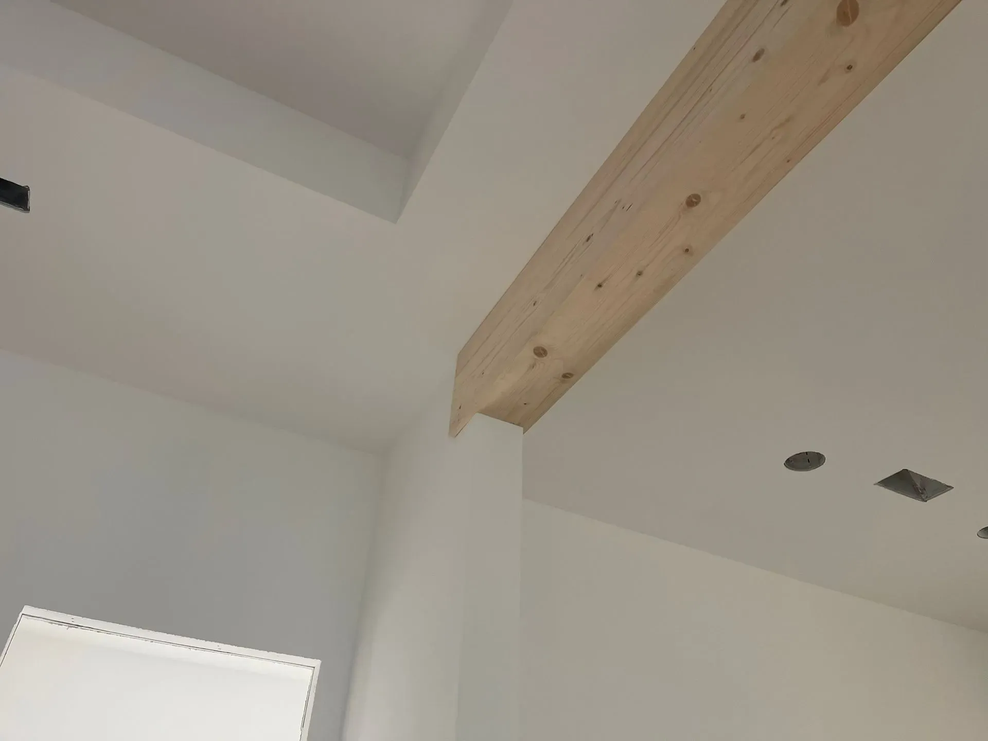 White ceiling with exposed wooden beam and recessed lights above a corner wall and window trim