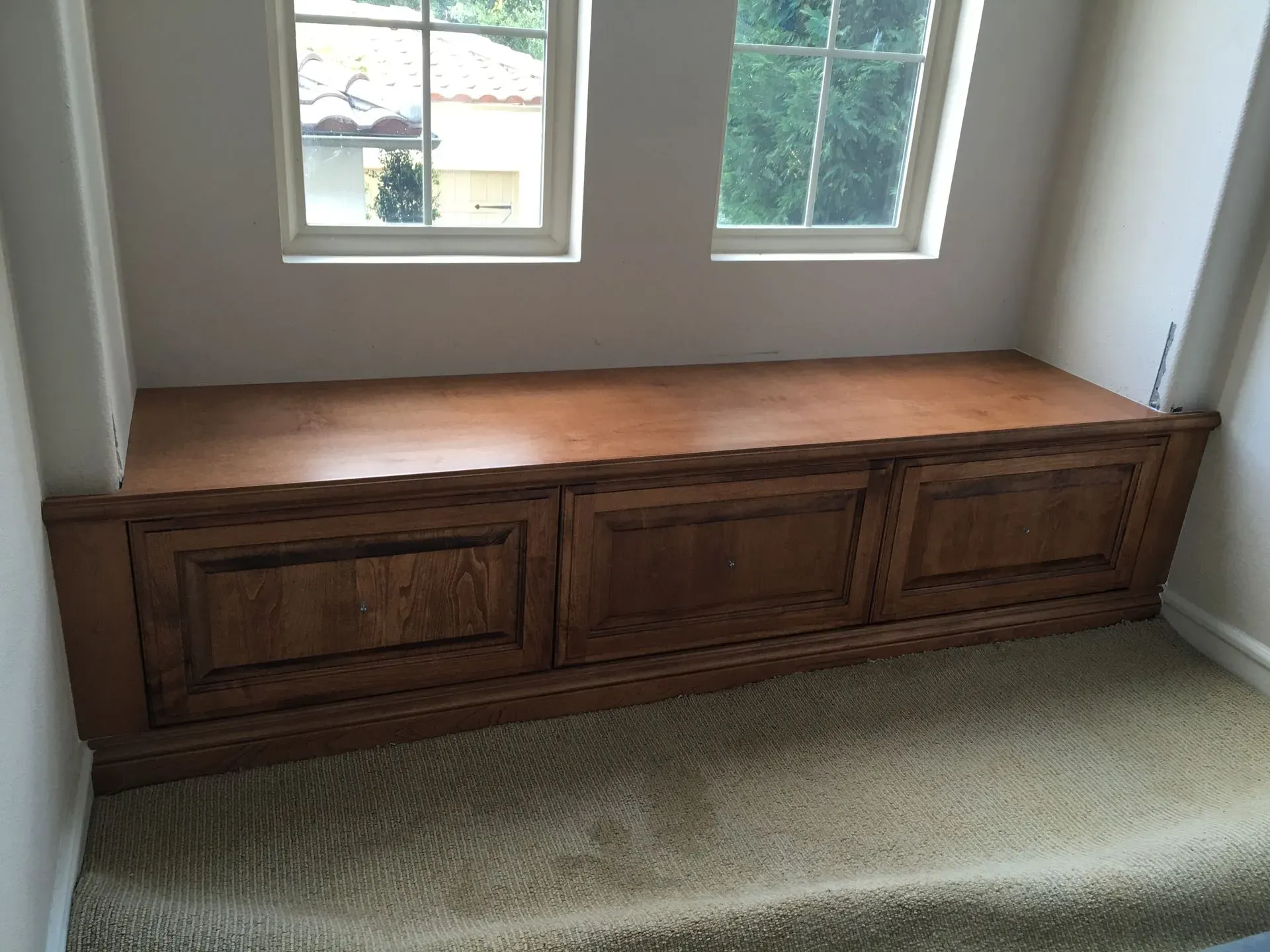Wooden storage bench under two windows in a carpeted room