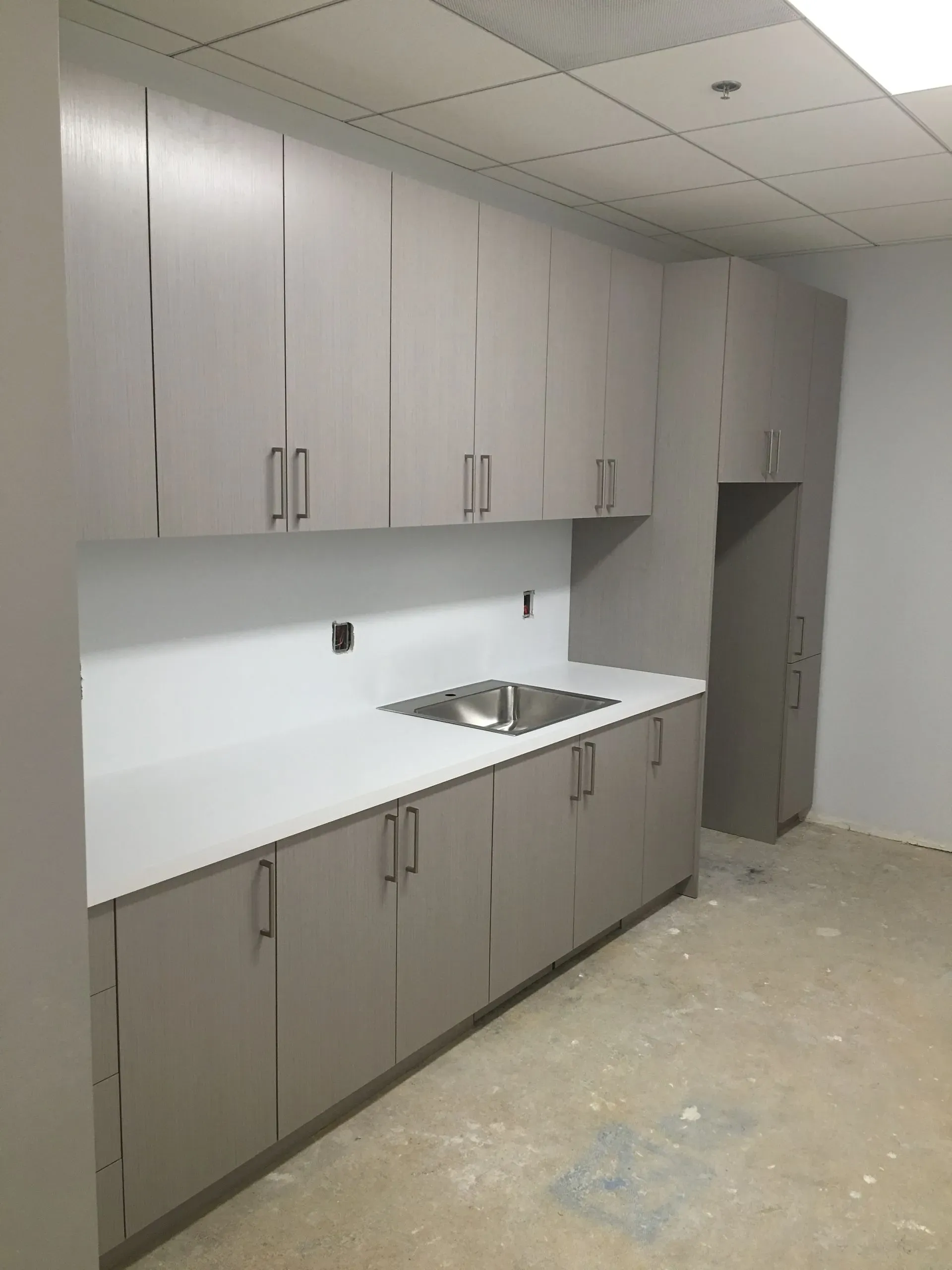 Empty kitchen with gray cabinets, white countertop, and stainless sink under fluorescent ceiling lights