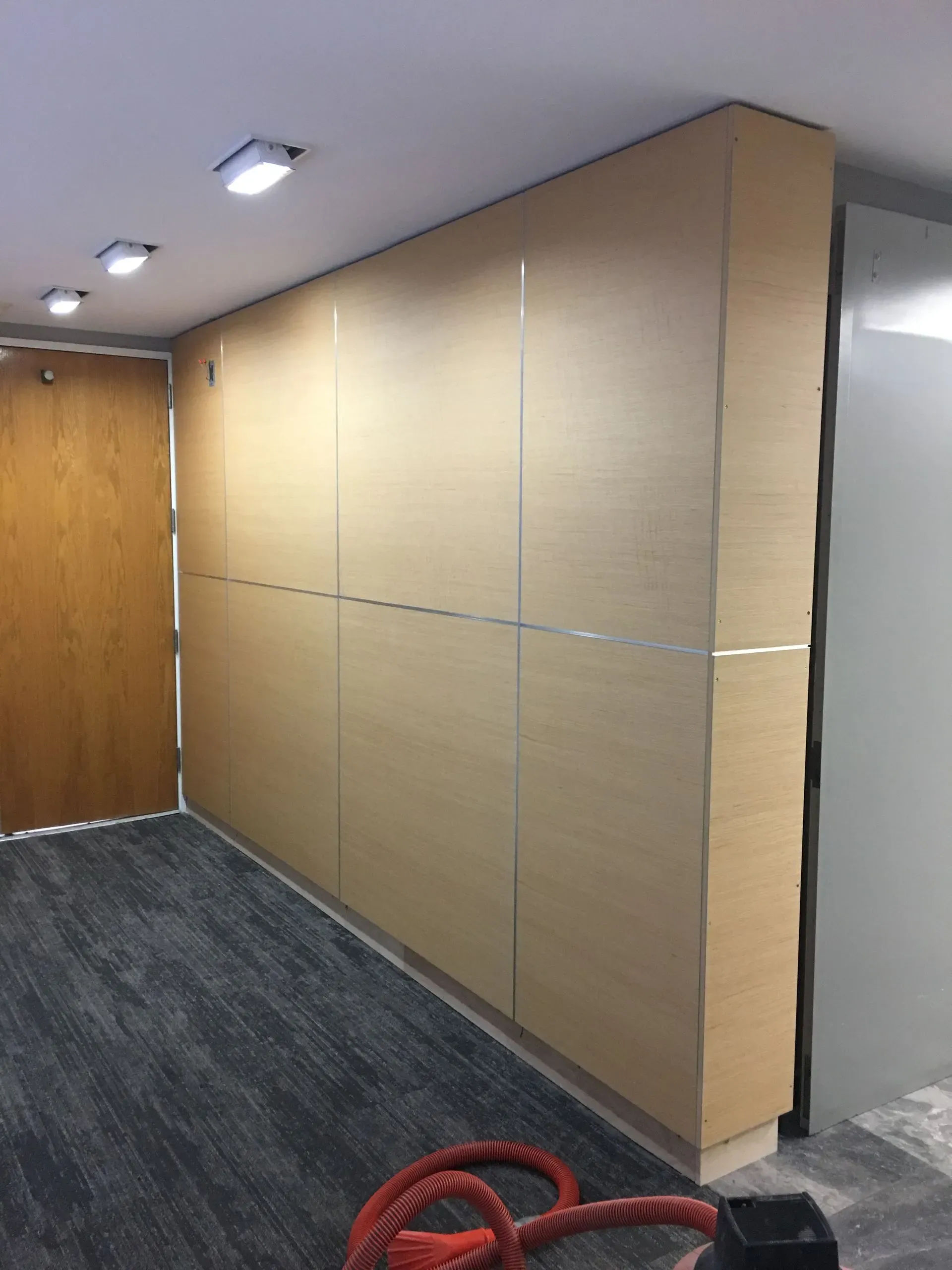 Hallway with tan wall panels, gray carpet, ceiling lights, and a red coiled cable on the floor.