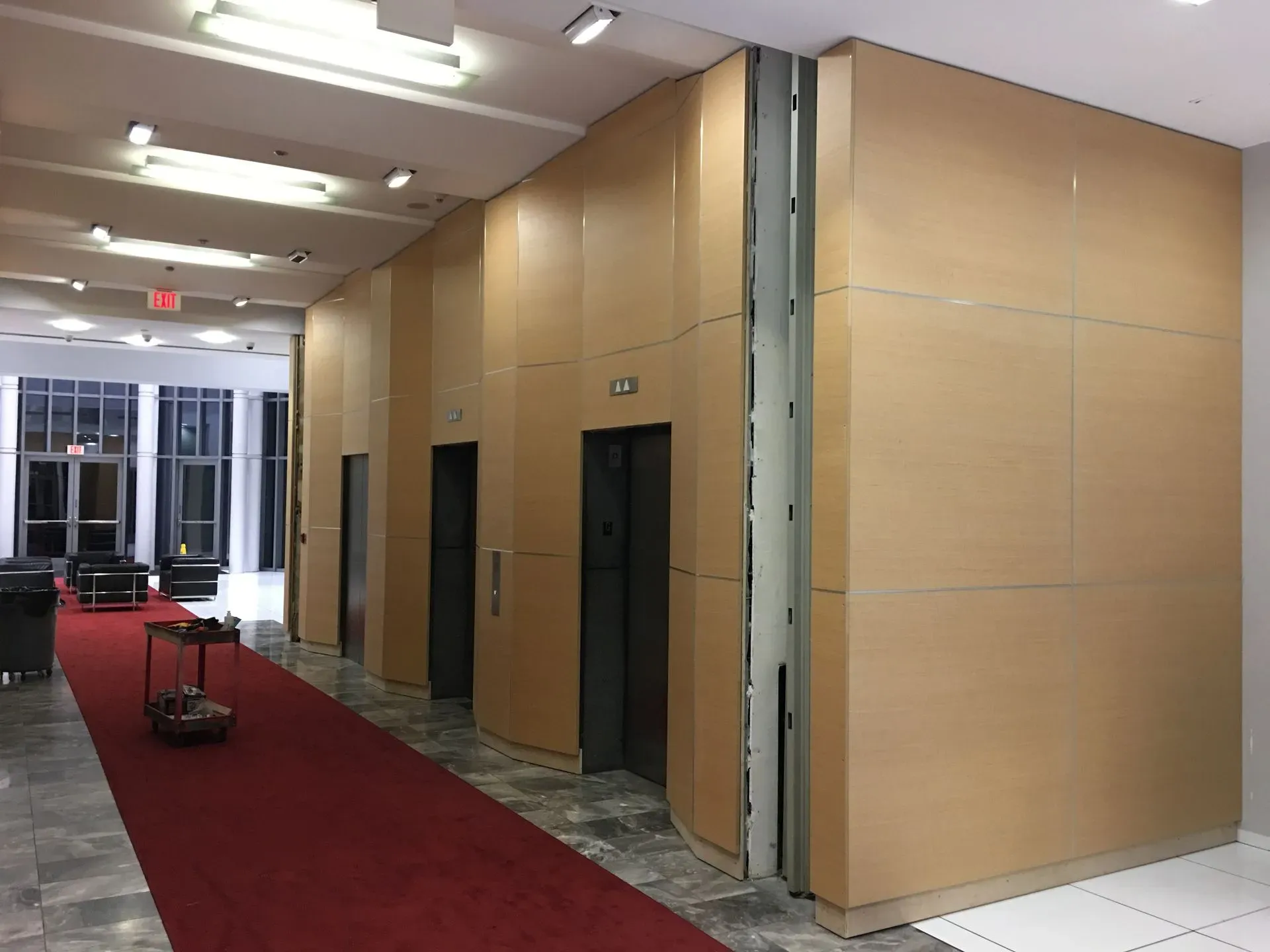 Empty hotel corridor with red carpet and wood-paneled walls, viewed toward glass doors.