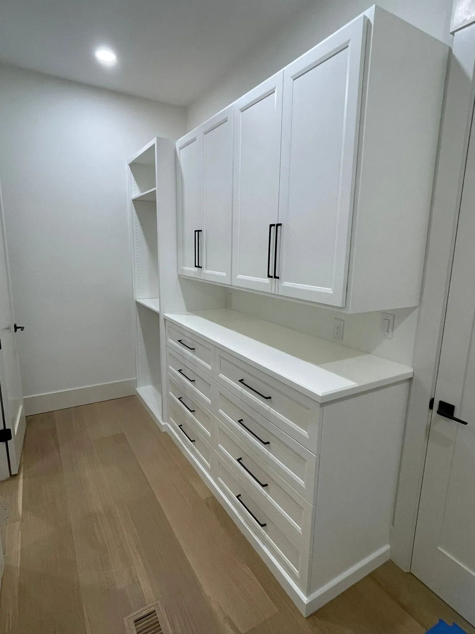 White hallway storage cabinets with drawers and upper cupboards in a narrow room with wood floor.