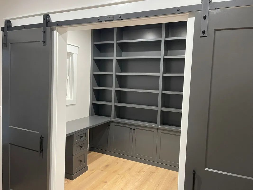 Open closet with gray shelving, built-in desk, and light wood floor, framed by dark barn doors