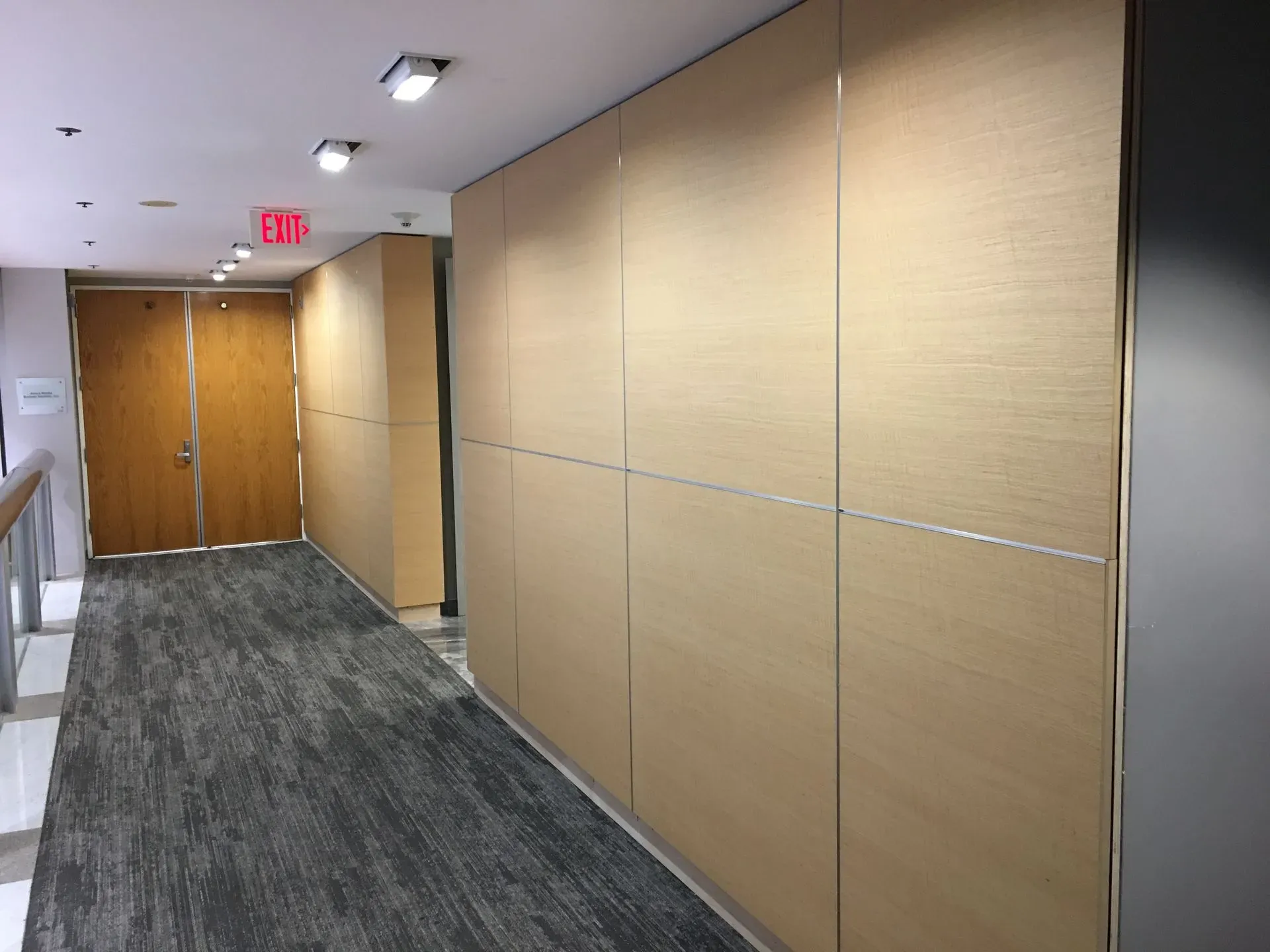 Empty office hallway with beige walls, gray carpet, and double wooden doors at the end.