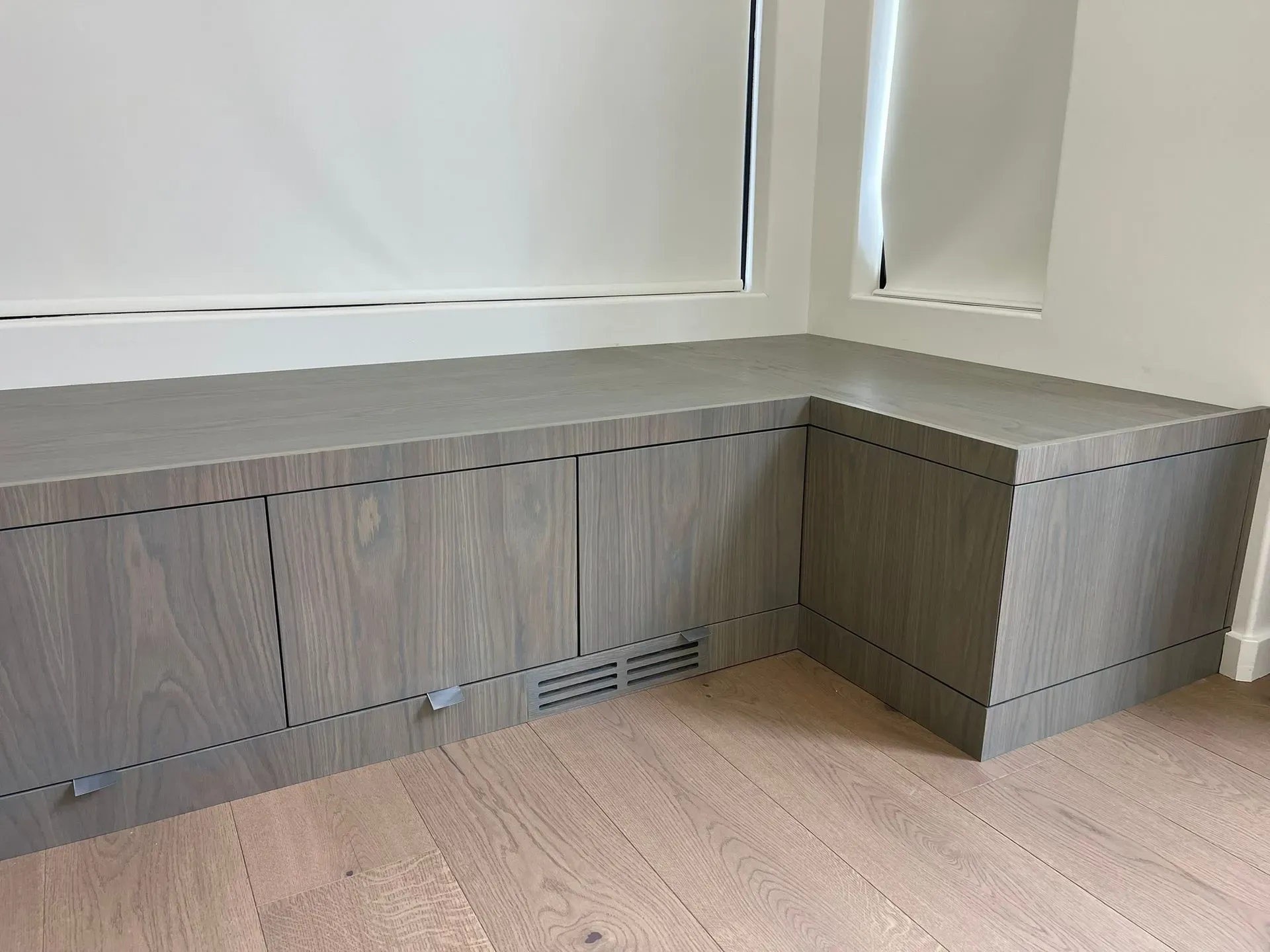Built-in gray storage cabinets under a window in a corner room with light wood flooring