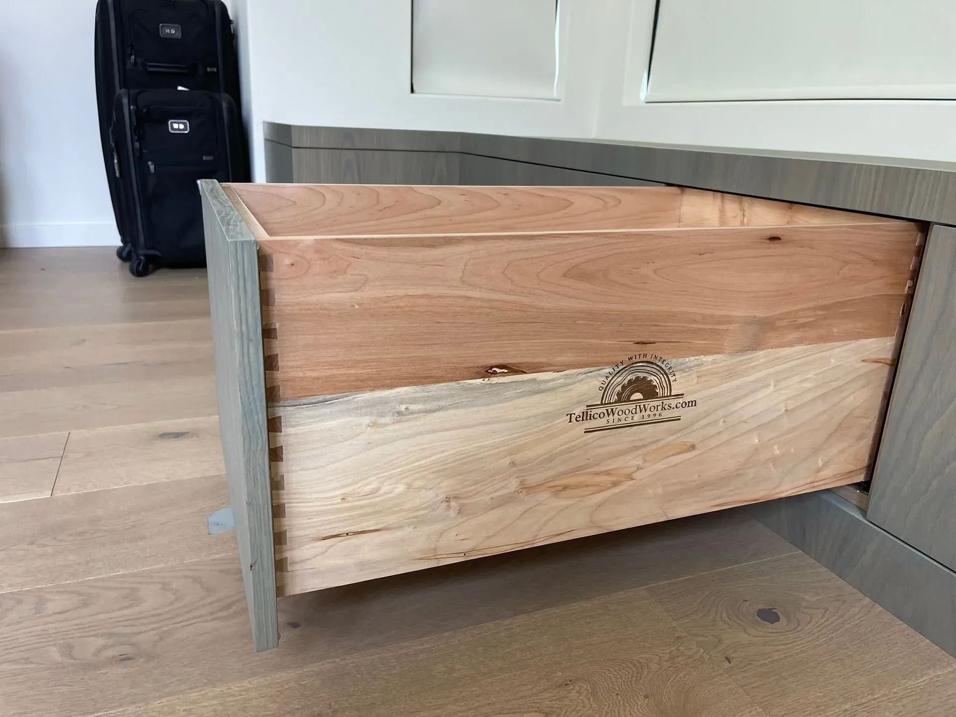 Open wooden drawer in a light wood kitchen cabinet with a suitcase in the background