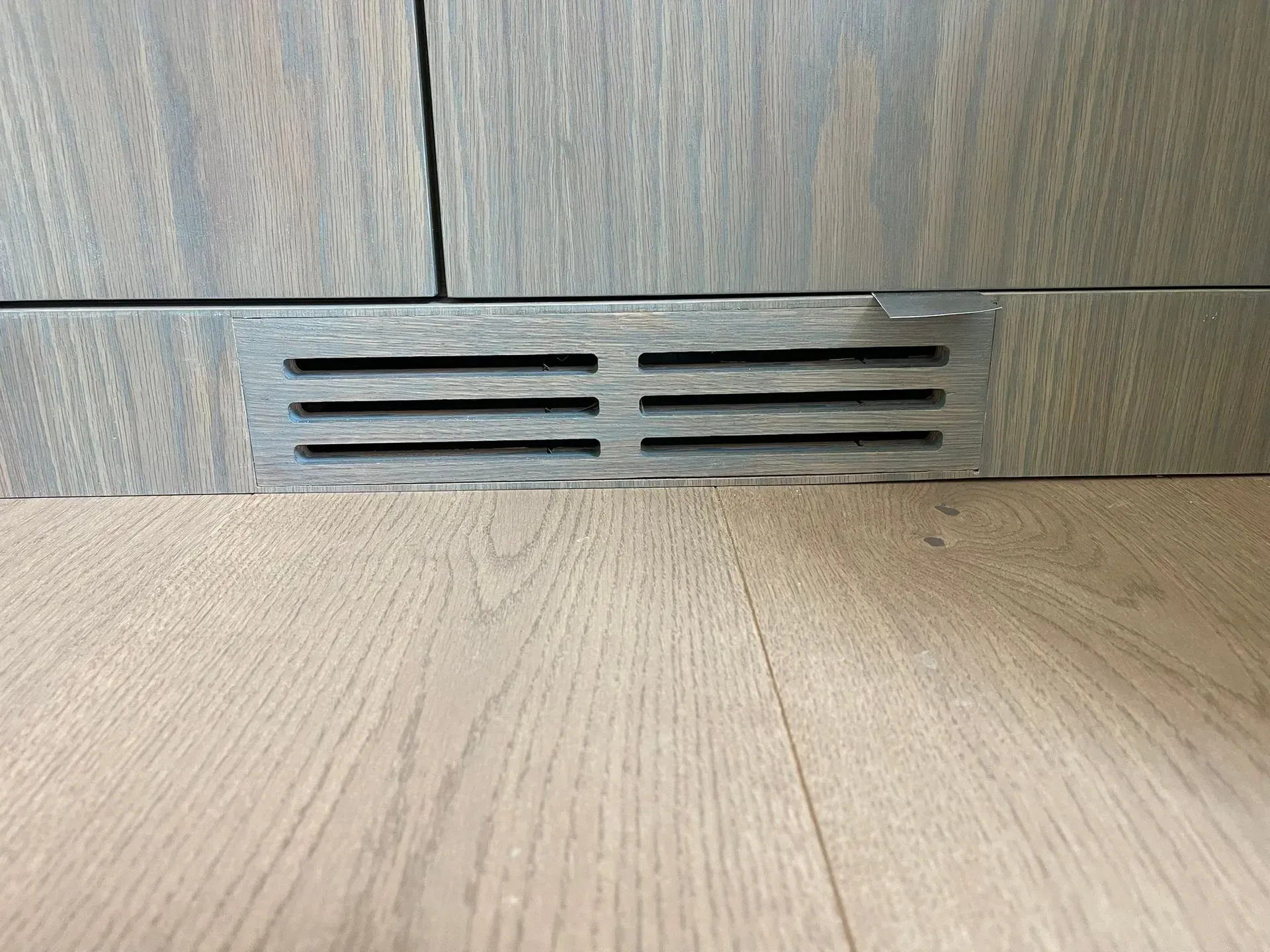 Close-up of a gray vent grille set into a wood-paneled wall above a light wood floor