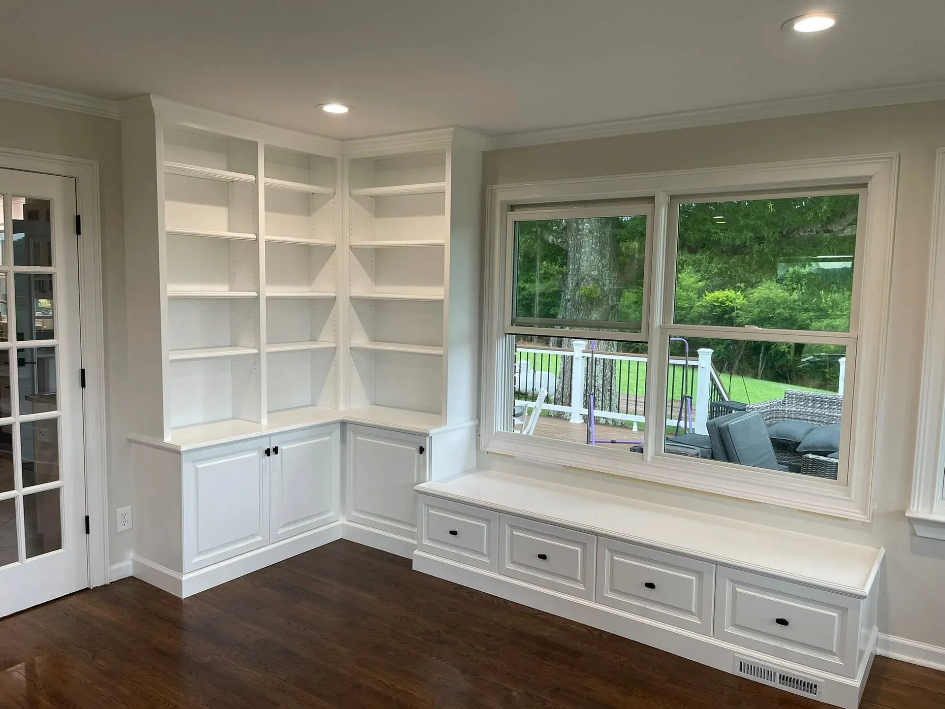Bright white built-in shelving and cabinets in a room with a large window and dark wood floor.