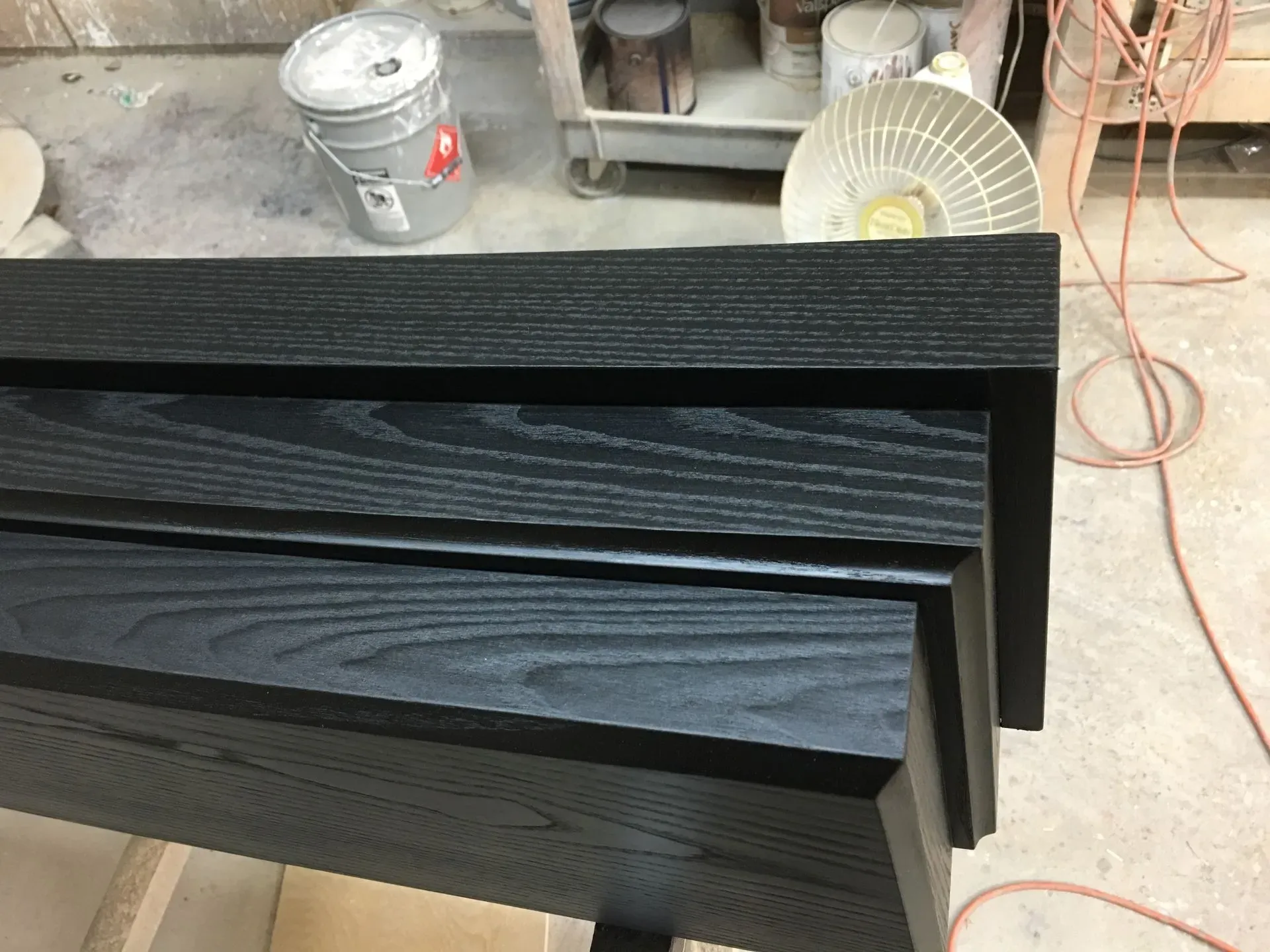 Stacked black wood planks in a workshop, with a bucket and fan in the background