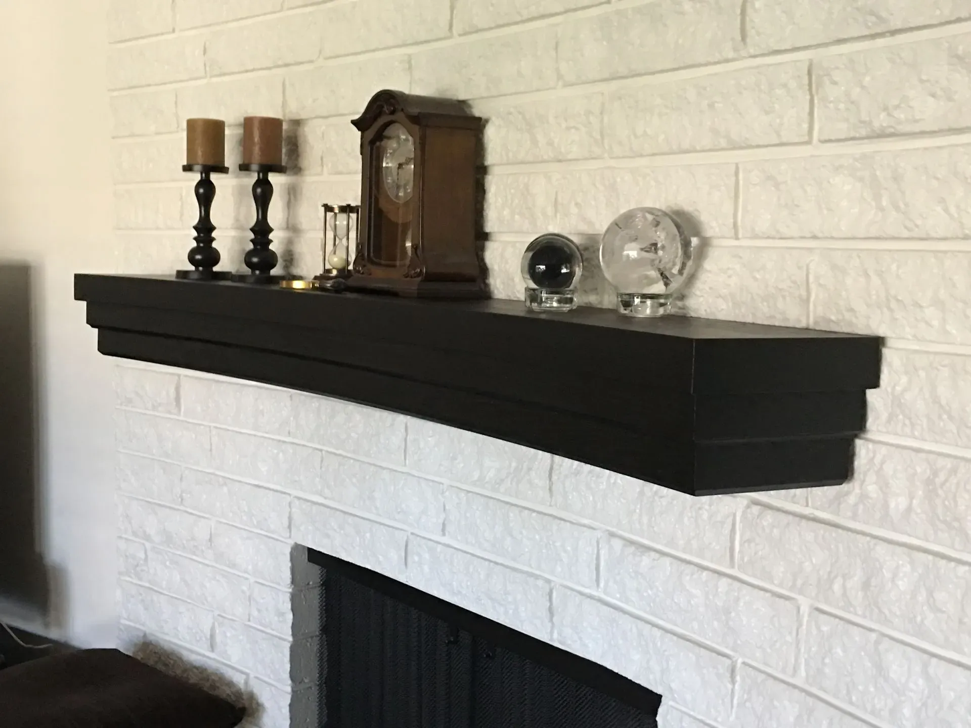 Black mantel shelf with candles, clock, and glass decor above a white brick fireplace