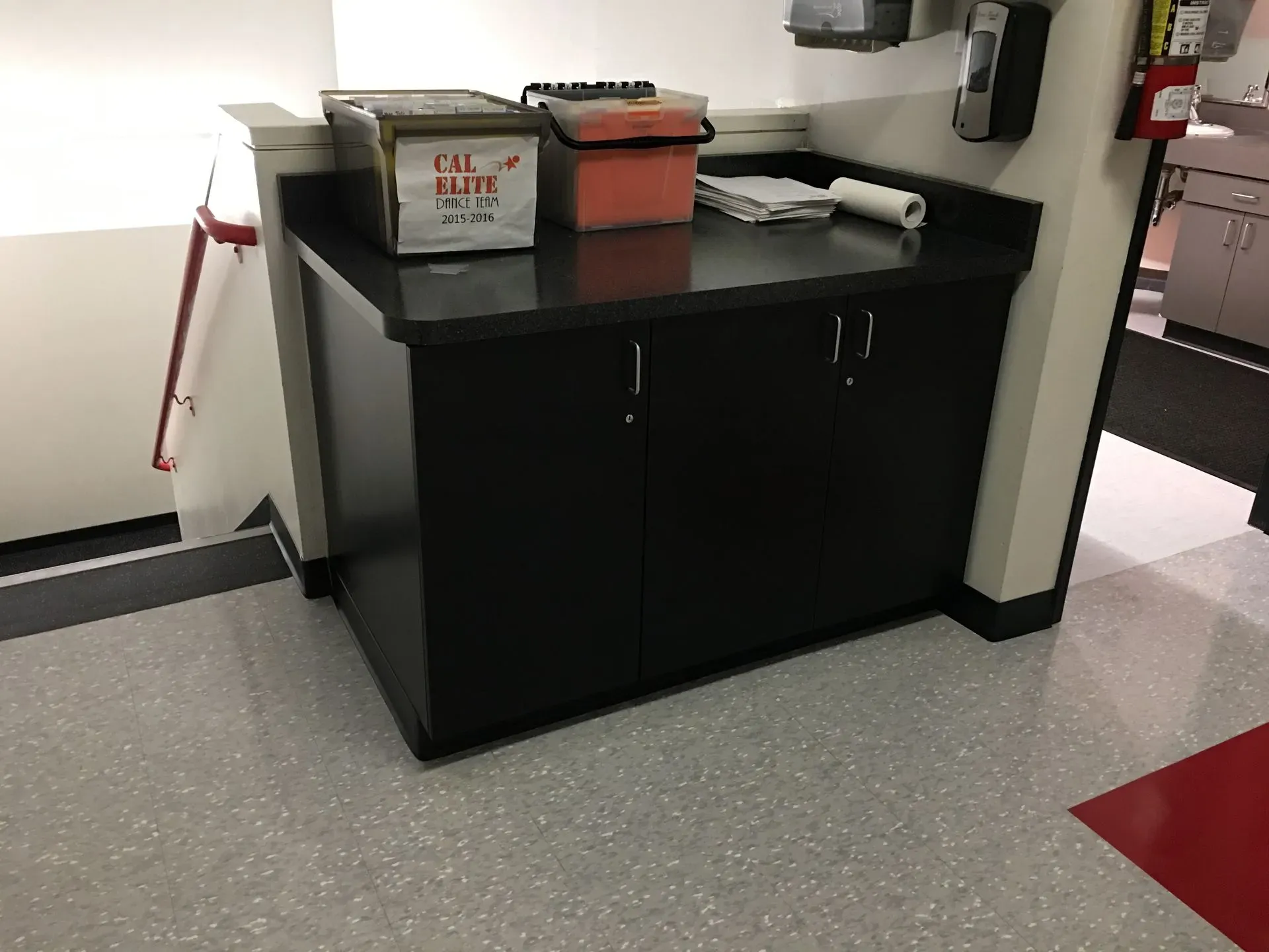 Black cabinet with boxes and supplies in a hallway or break room corner.