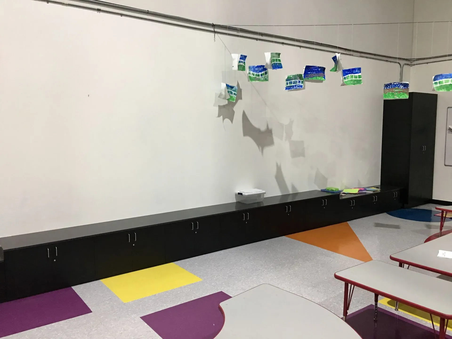Empty classroom with whiteboard, colorful floor mats, hanging decorations, and tables and chairs along the wall