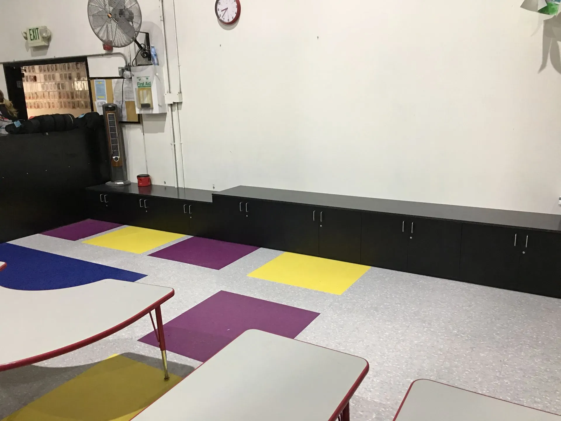Empty classroom with colorful foam floor tiles, black storage cabinets, and a wall fan.