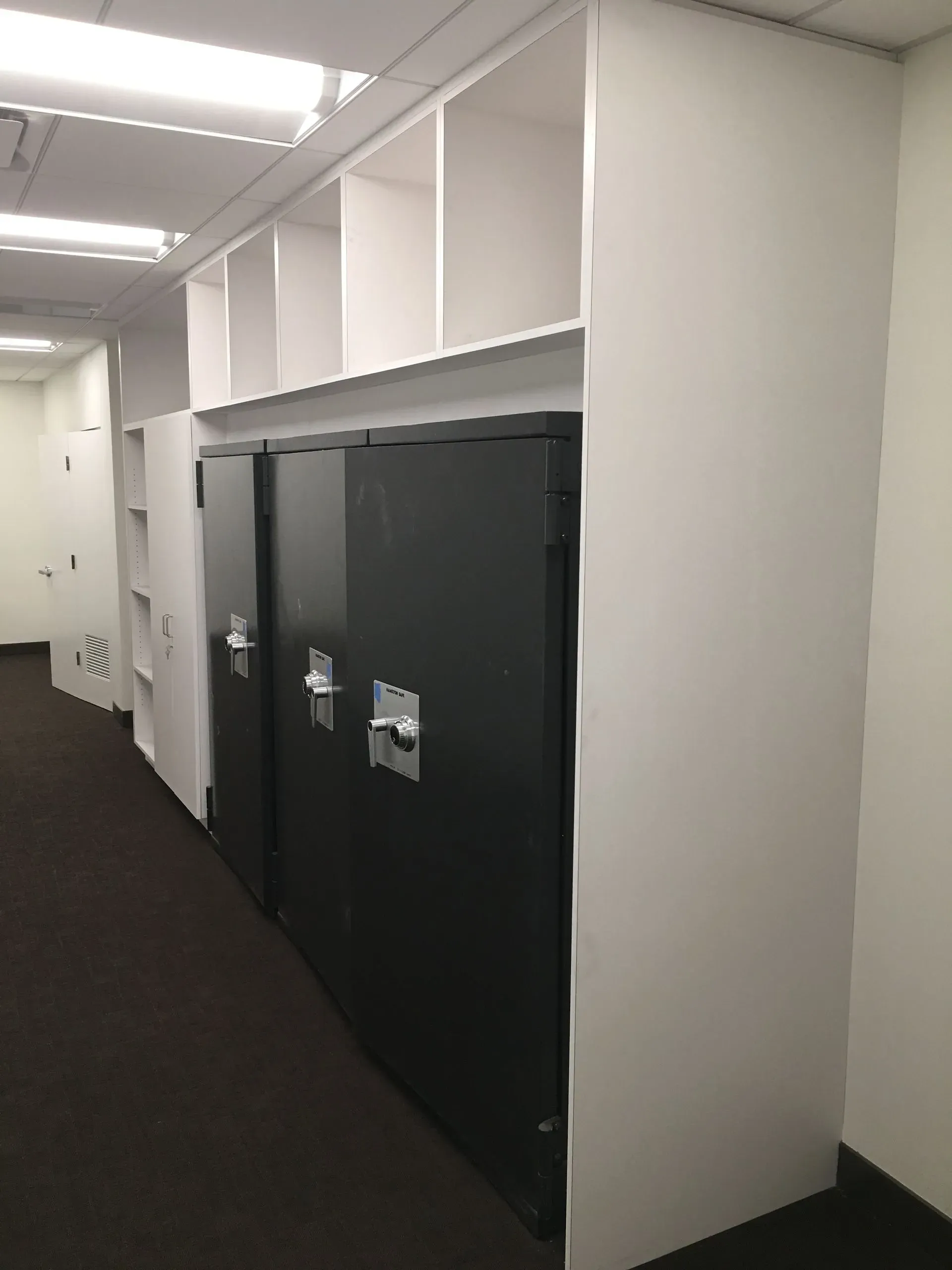 Office hallway with dark storage lockers along a white wall under fluorescent ceiling lights