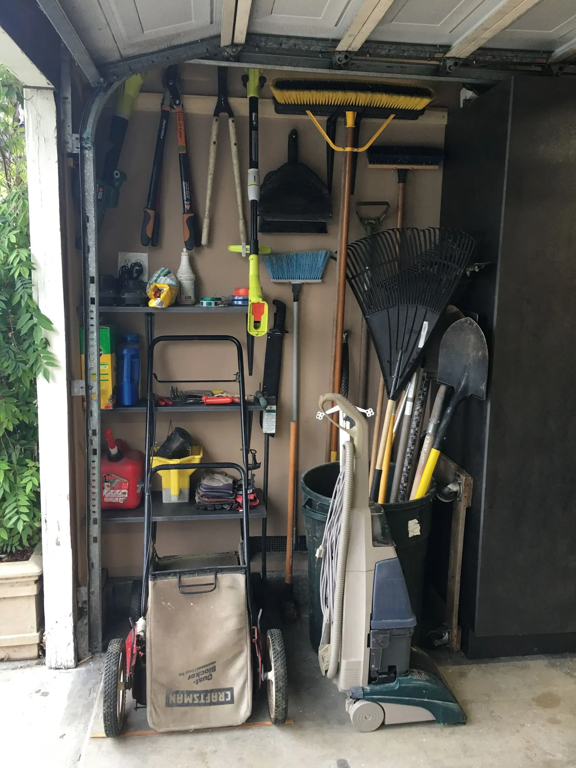 Garage storage area with shelves, tools, rakes, and a lawn mower in a narrow cluttered space