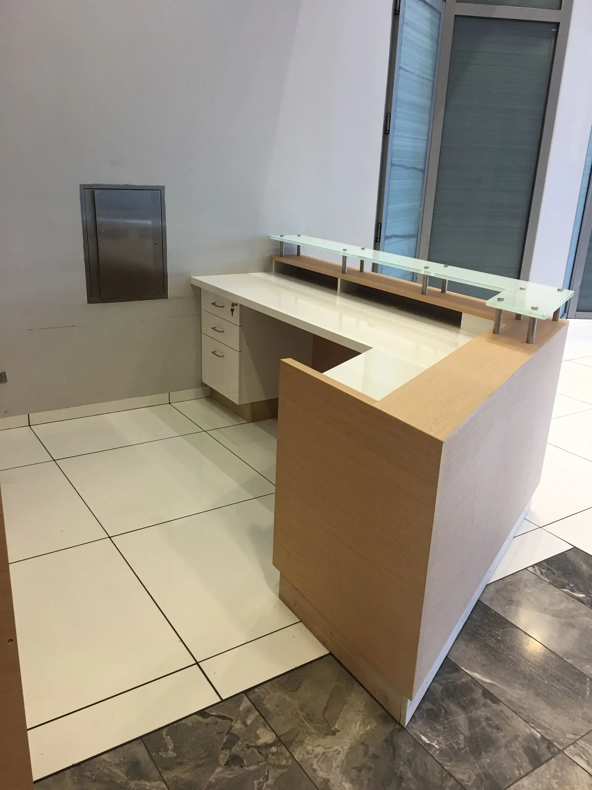 Empty reception desk with white and wood counter in a tiled lobby, beside a wall-mounted dispenser.
