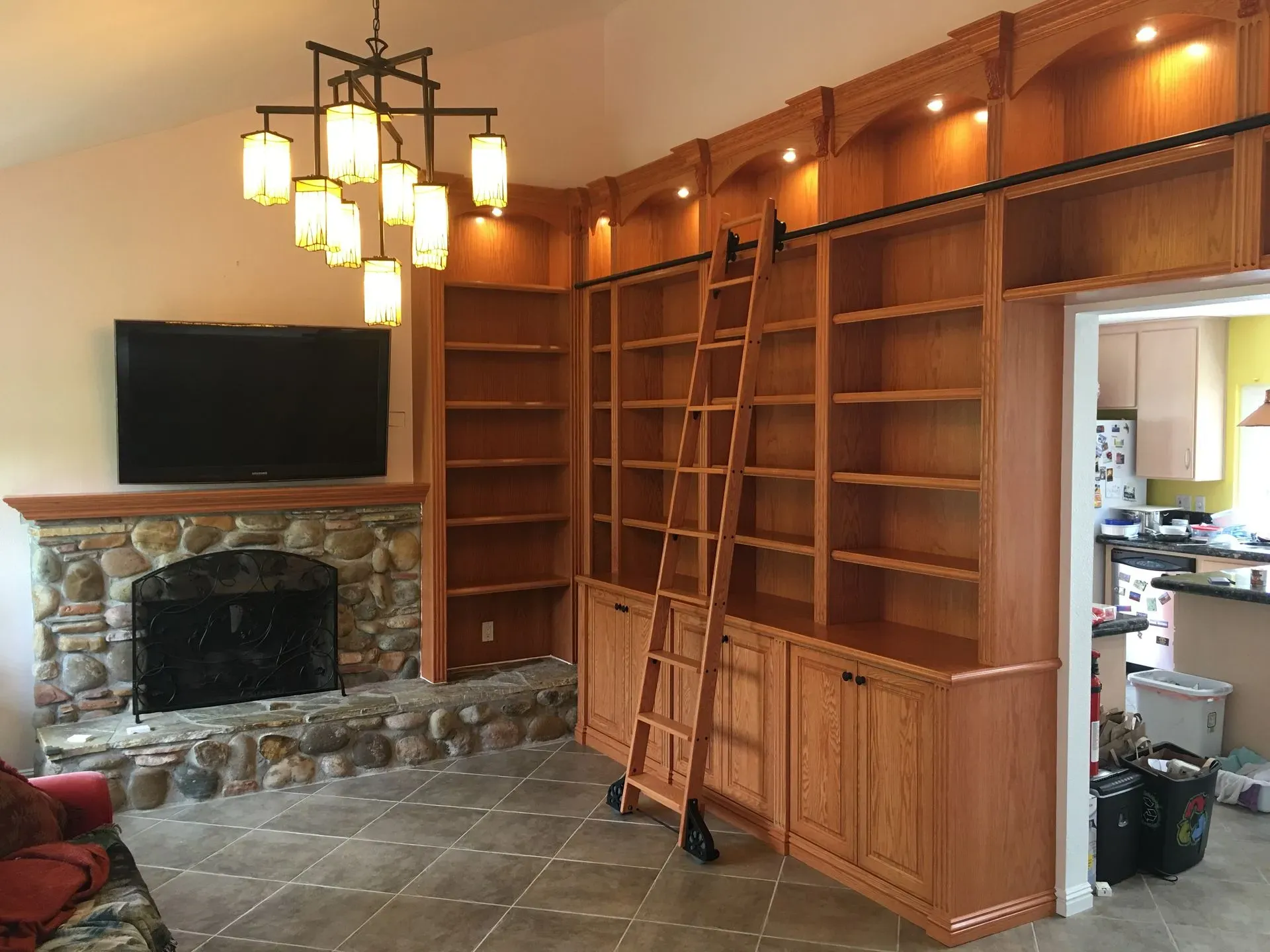 Wood-paneled living room with stone fireplace, built-in bookshelves, TV, and ladder under warm ceiling lights