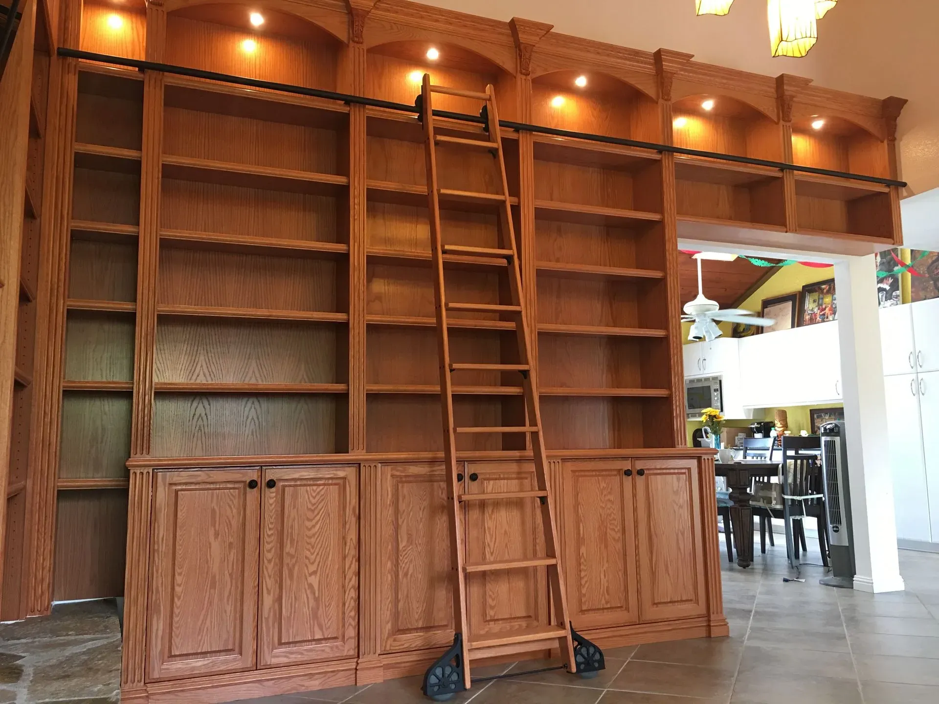 Wooden bookshelf wall with a central ladder in a warmly lit room, open doorway to a café area