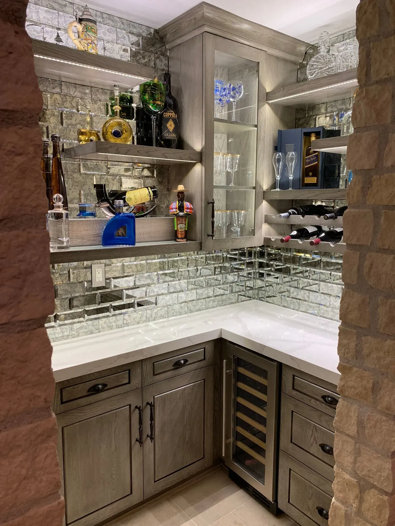 Built-in corner bar with mirrored shelves, glassware, bottles, and white countertops in a home interior