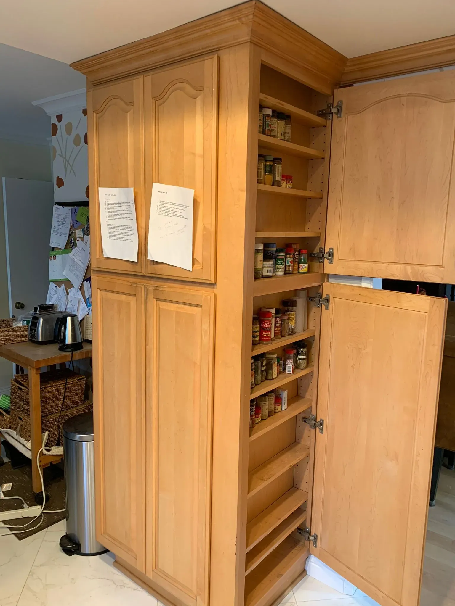 Tall wooden kitchen pantry cabinet with pull-out shelves and two notes posted on the front doors.