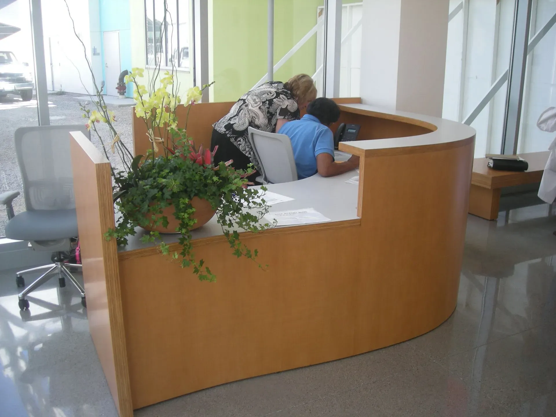 Two people leaning over a curved wooden reception desk in a bright office lobby