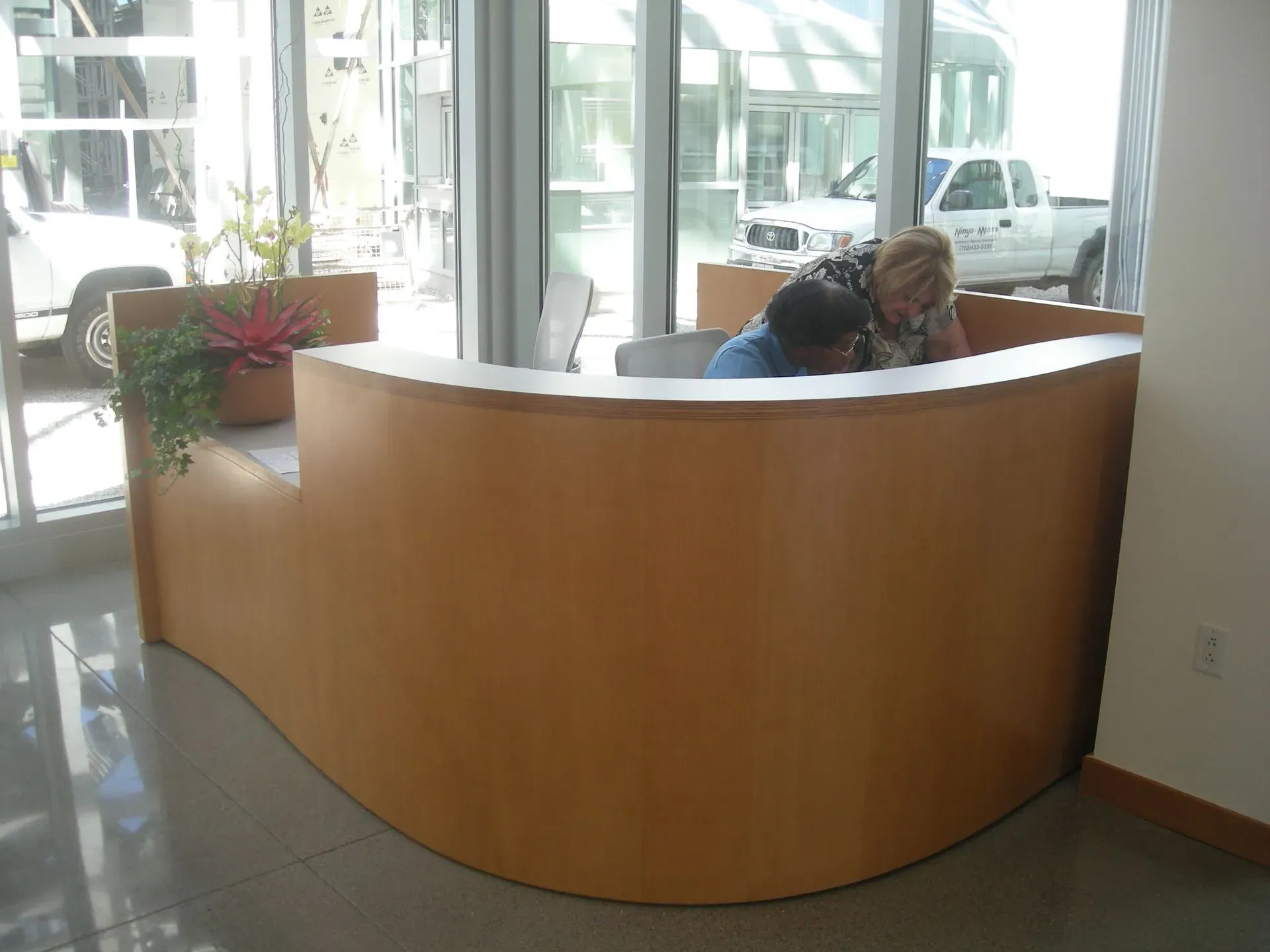 Curved wooden reception desk in a bright lobby, with a person seated behind it near large windows.
