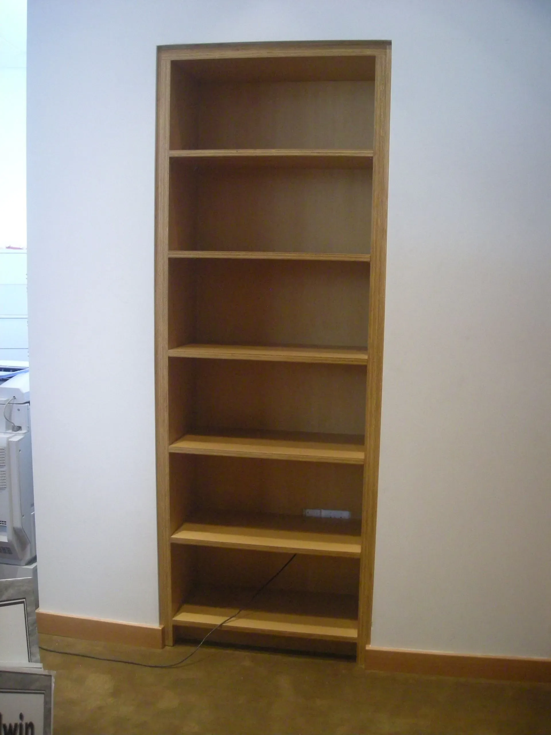 Tall wooden bookshelf against a white wall on a wooden floor, with several empty shelves.