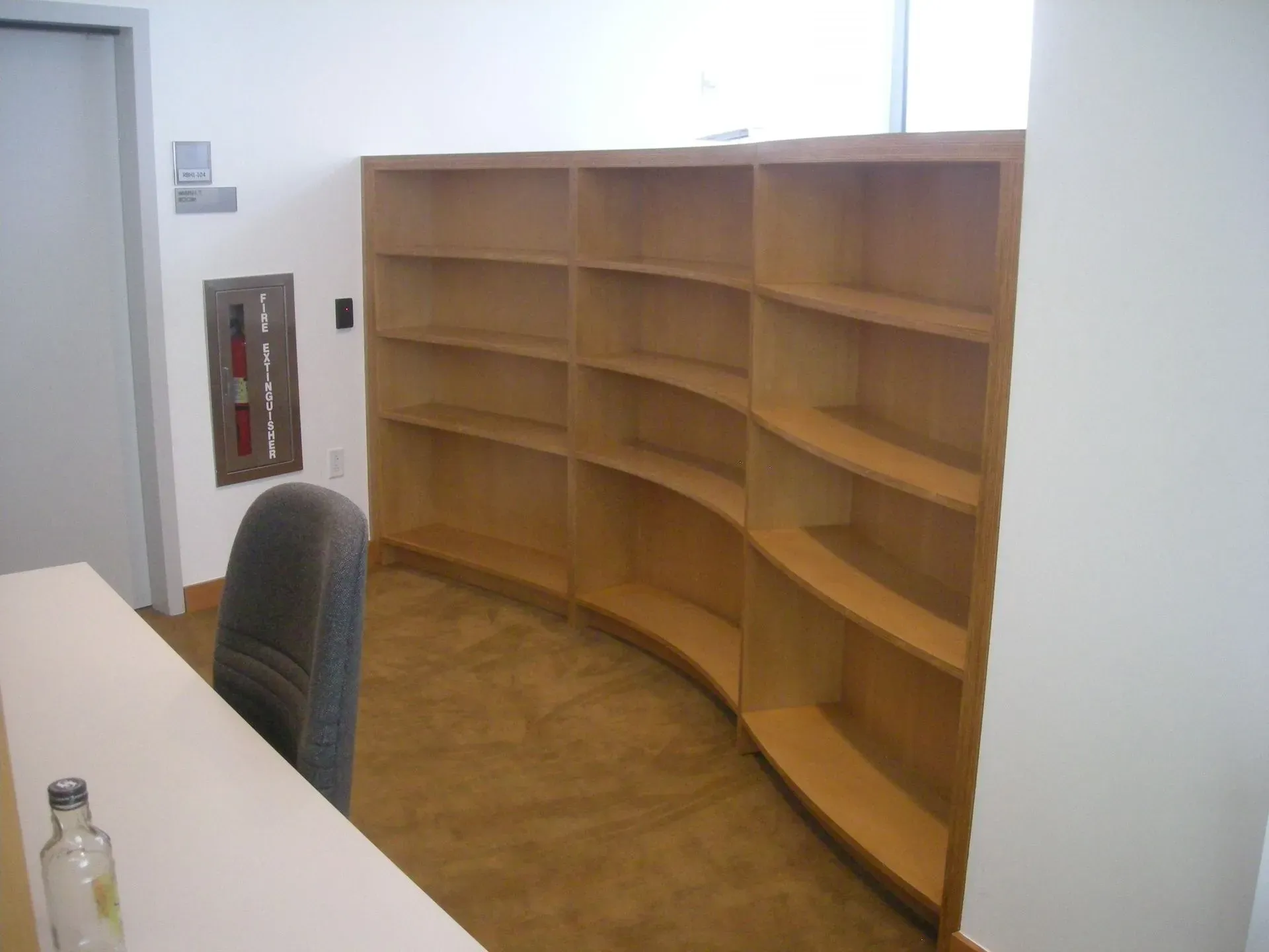 Empty office with curved wooden bookshelves, desk edge, chair, and fire extinguisher cabinet on the wall