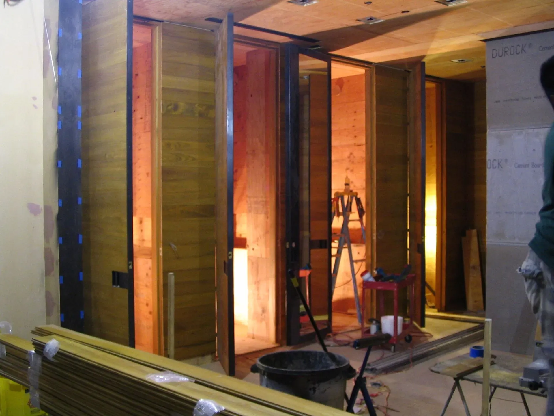 Interior construction site with exposed wooden framing, open wall panels, tools, and a ladder under warm lighting