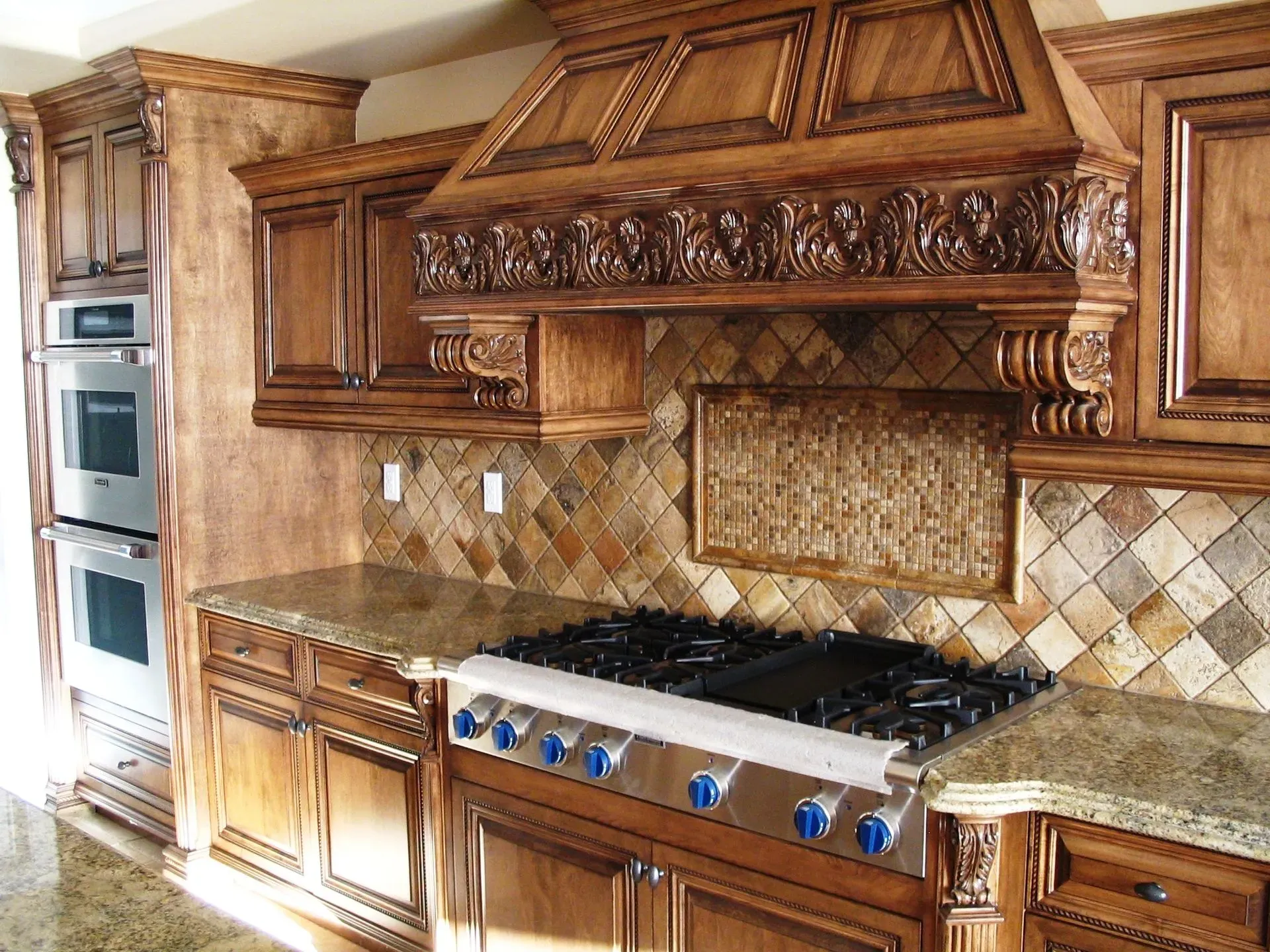 Ornate wooden kitchen with stainless steel stove and built-in ovens, tiled backsplash, and marble countertops