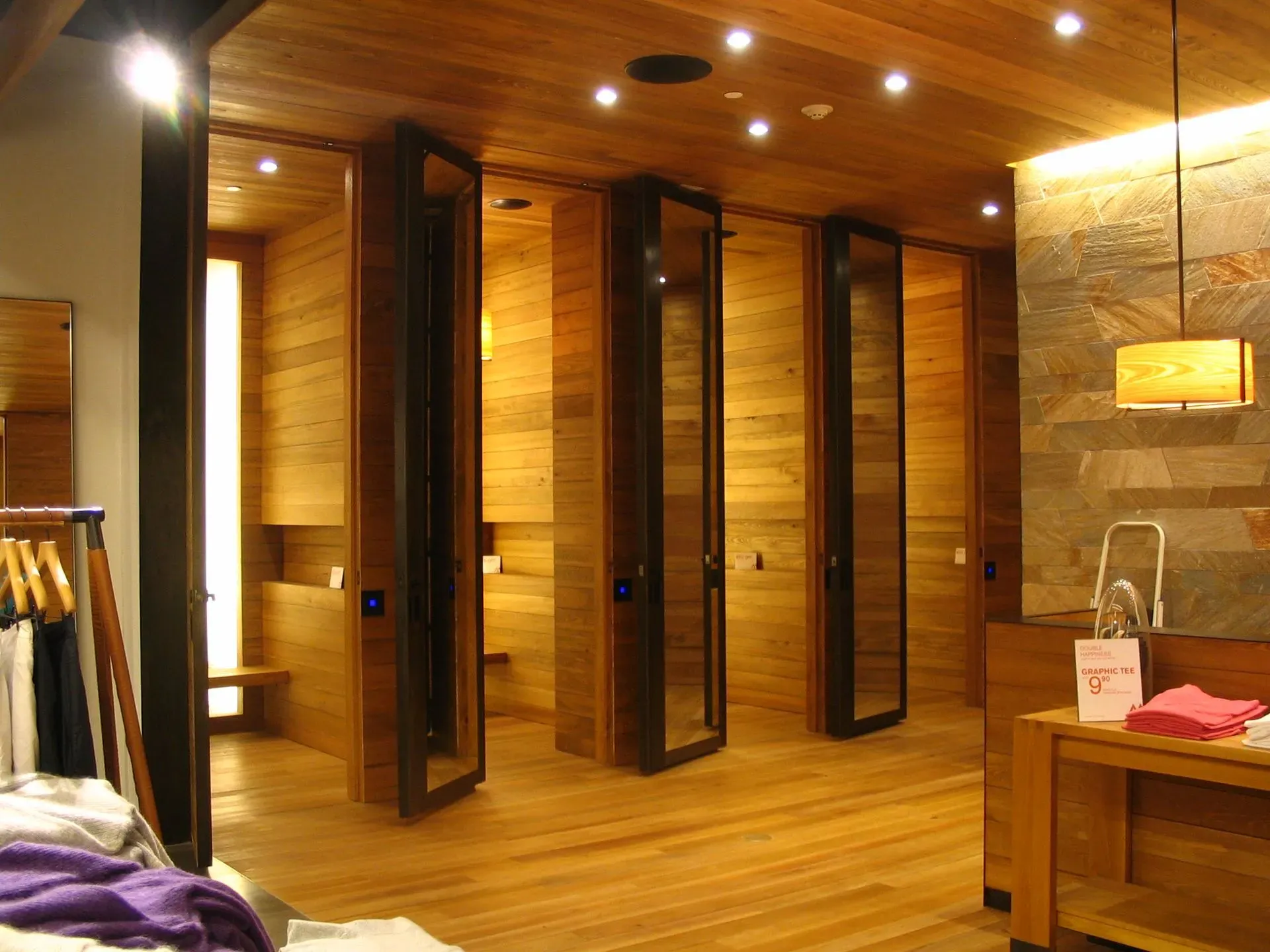 Wood-paneled fitting room with mirrored stalls, warm lighting, and clothing racks nearby