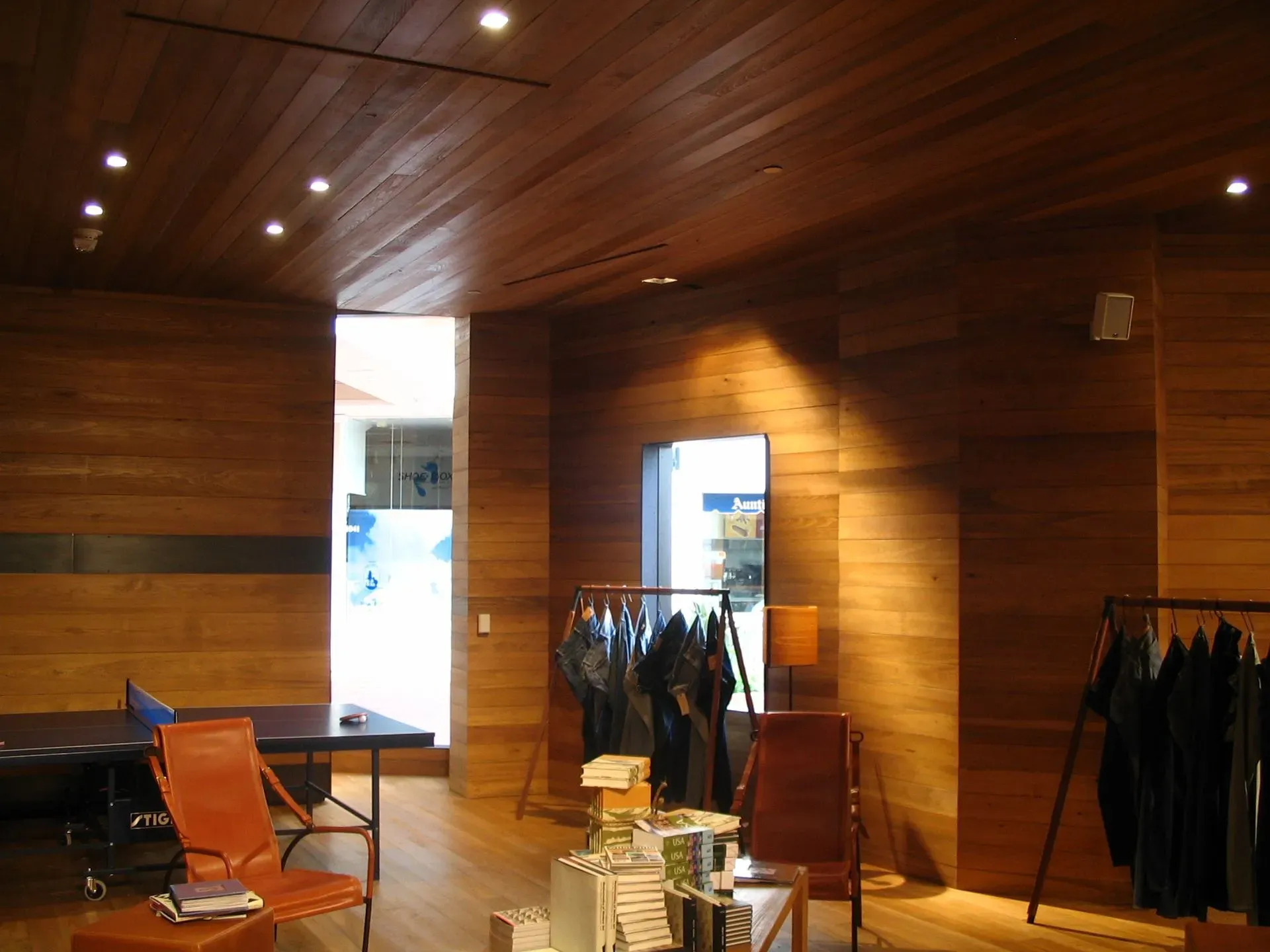 Wood-paneled clothing store interior with racks of dark clothes, a chair, and bright doorway windows