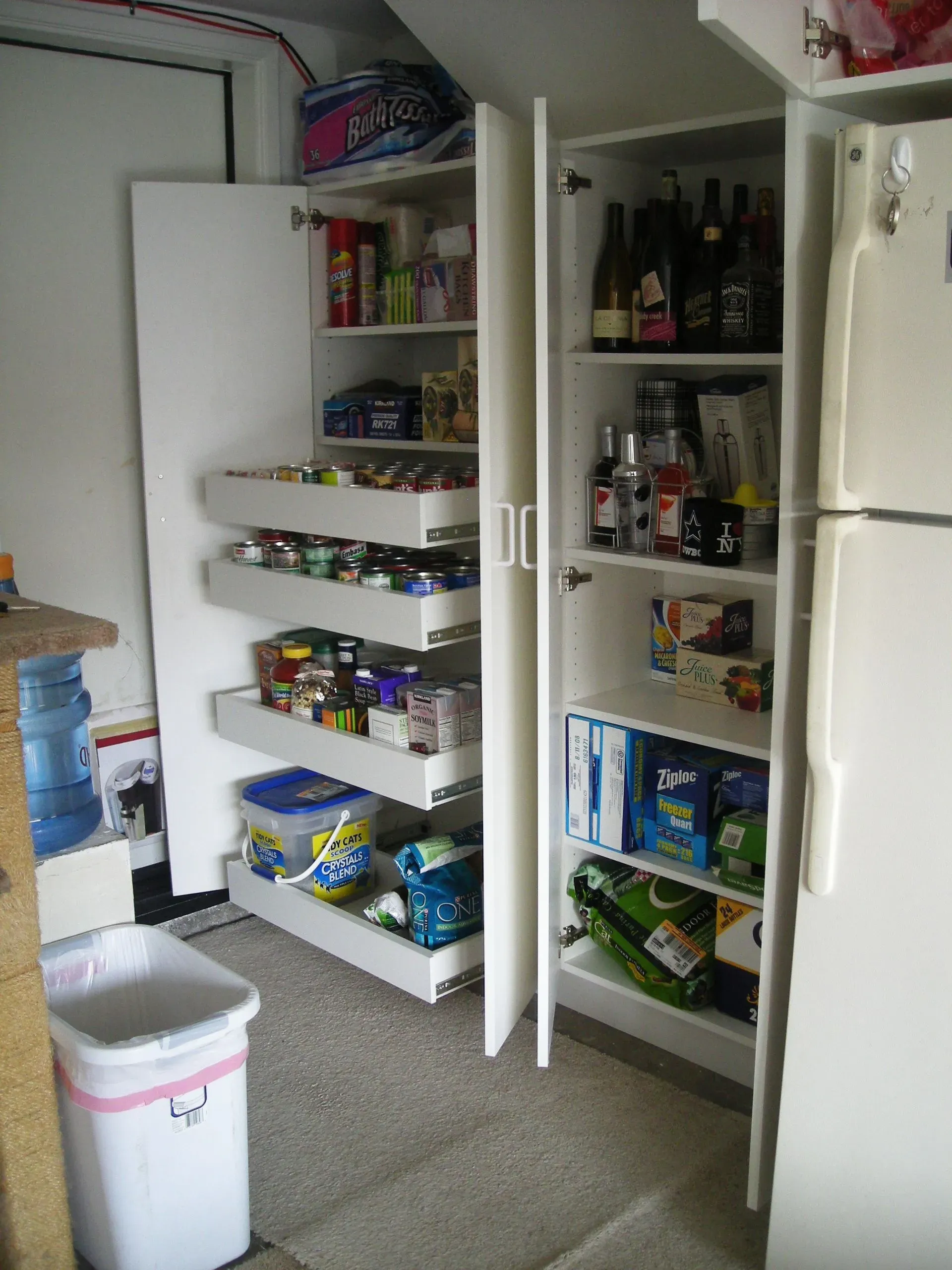 Open pantry with shelves of canned and boxed food beside a white refrigerator and trash bin
