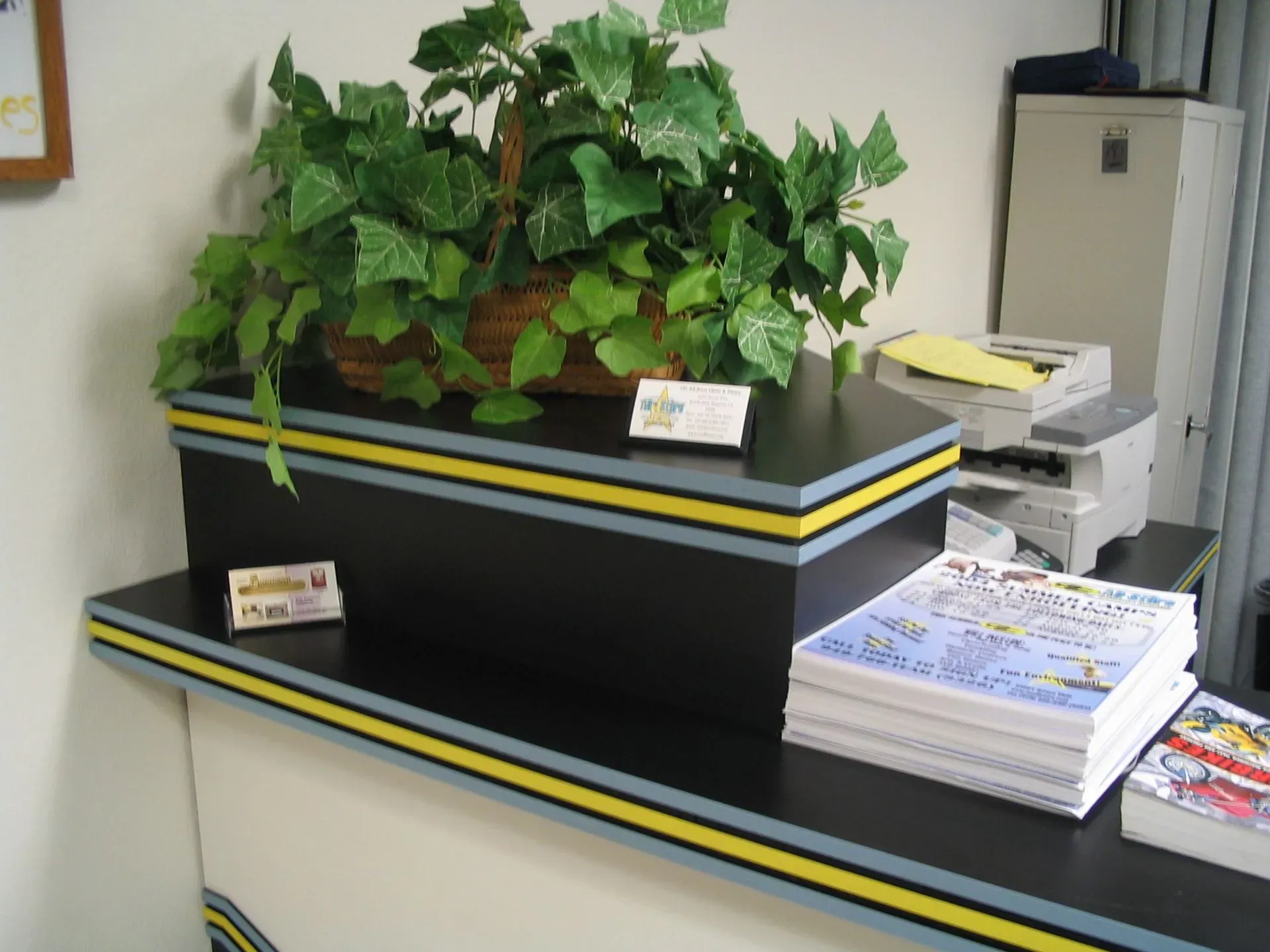 Office counter with a potted plant, stacked papers, and a printer beside a bulletin board