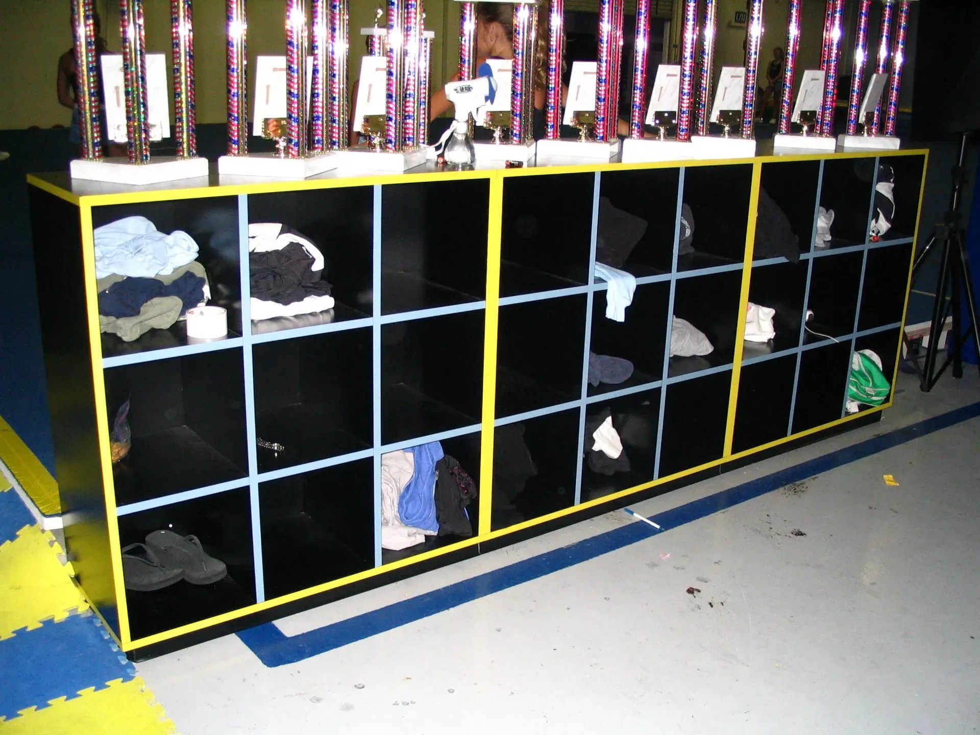Yellow cubby shelf with black bins and metal cups on top in a classroom or locker room area