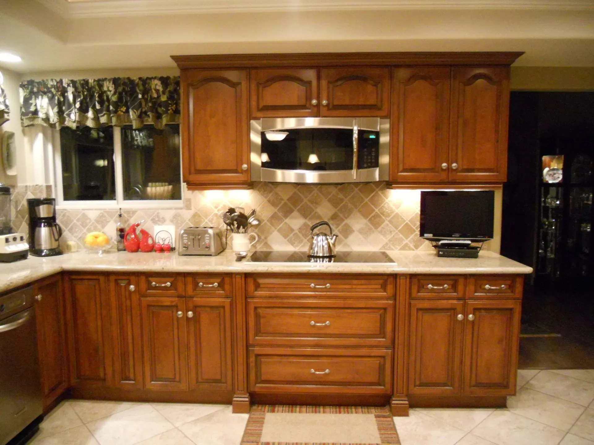 Wooden kitchen with tan tile backsplash, stainless microwave, countertop appliances, and dark doorway.