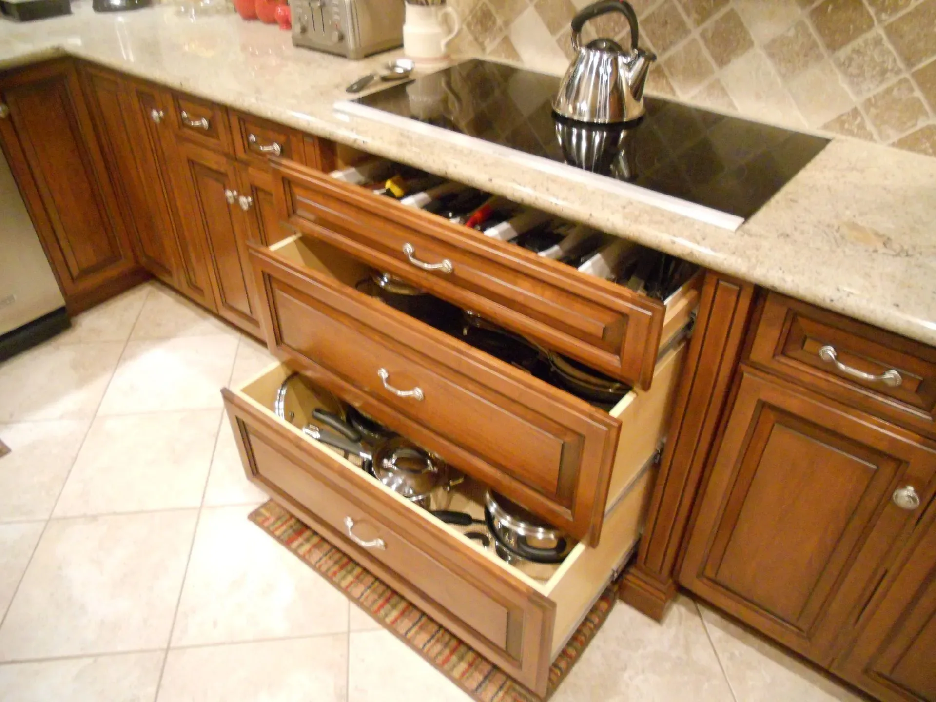 Kitchen cabinets with a drawer organizer pulled open, showing utensils and pots under a granite countertop.