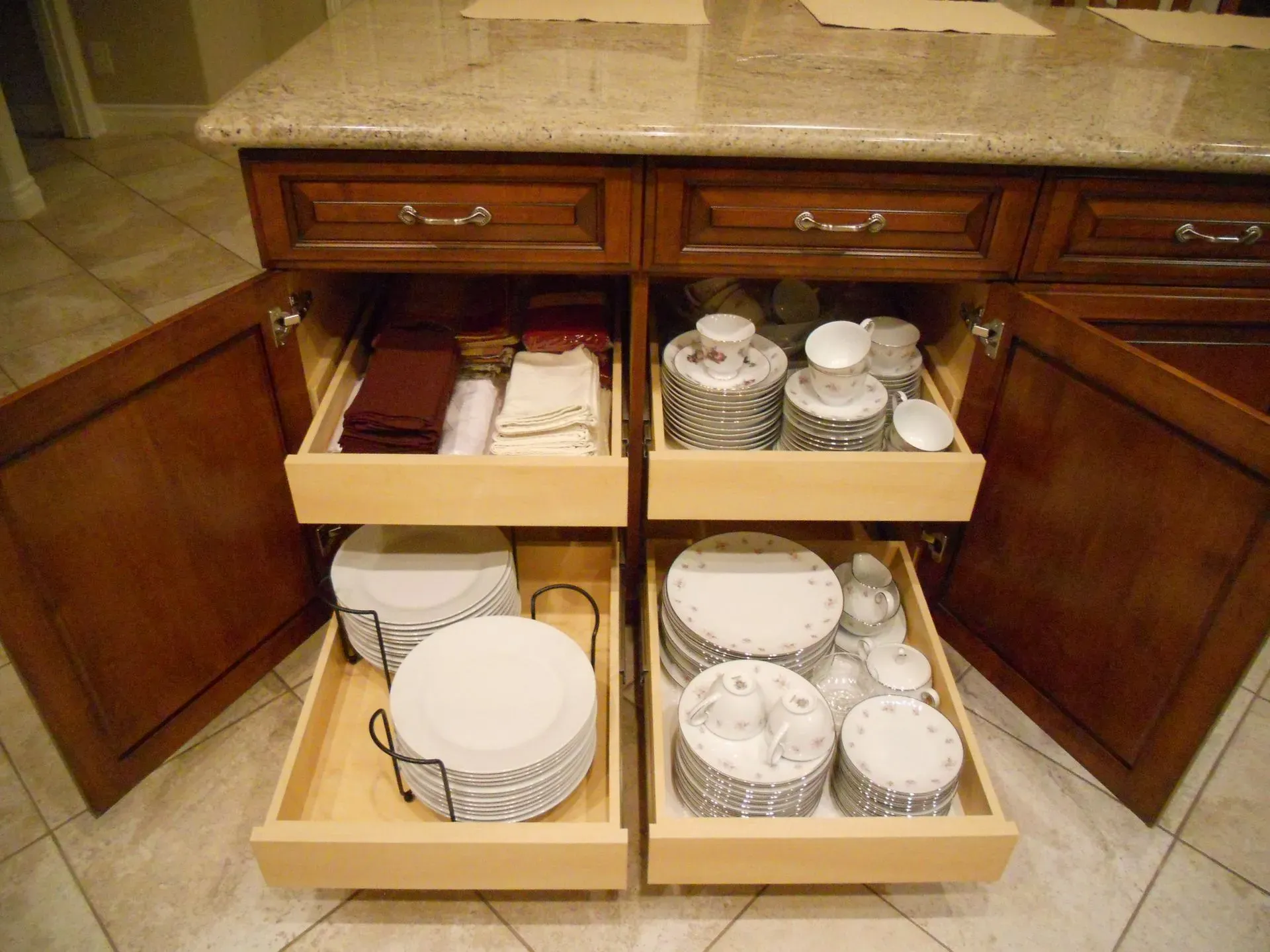 Open kitchen cabinet drawers holding dishes, cups, and utensils beneath a granite countertop