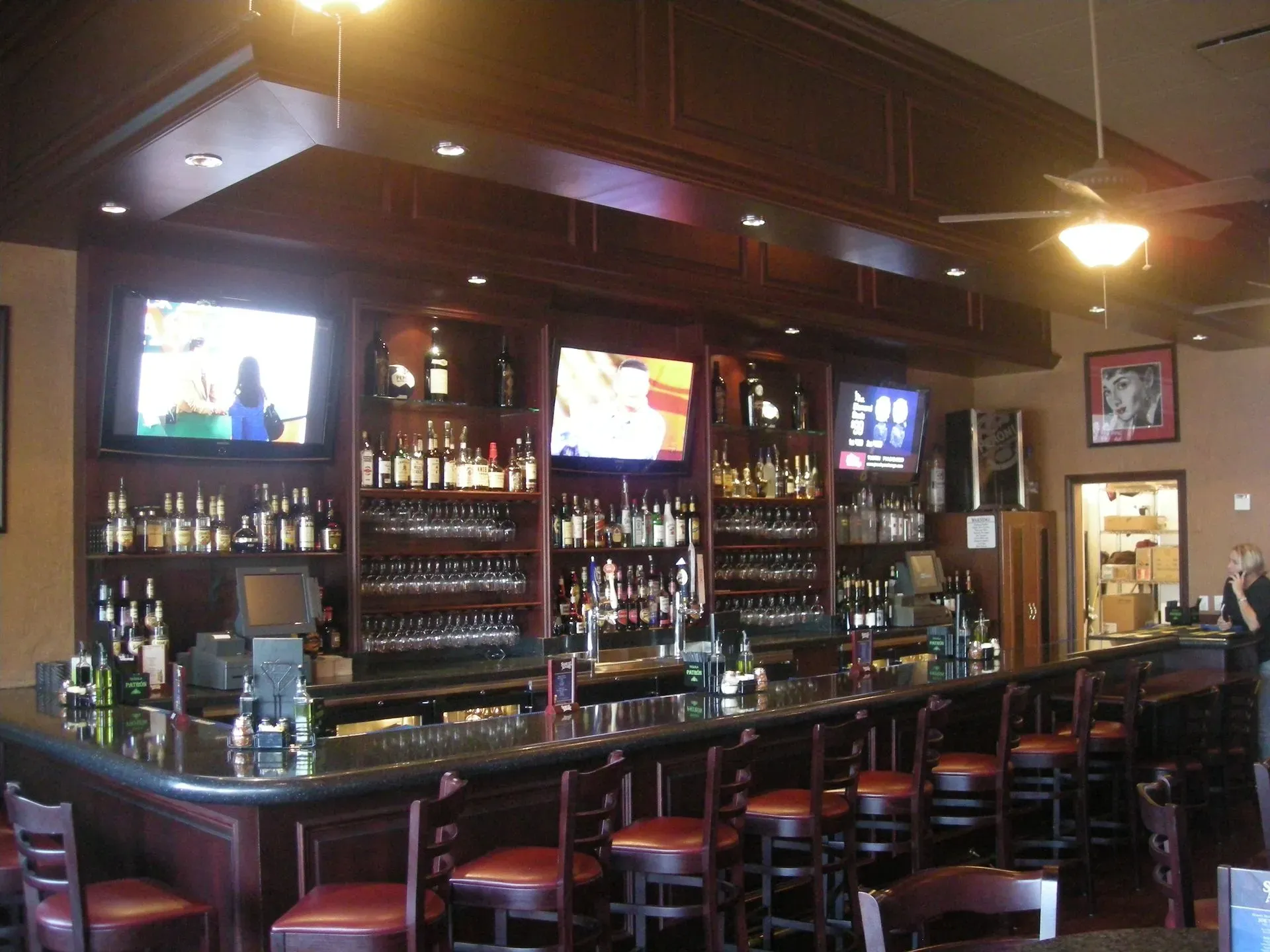 Dimly lit bar with liquor shelves, TV screens, and stools along a long counter.