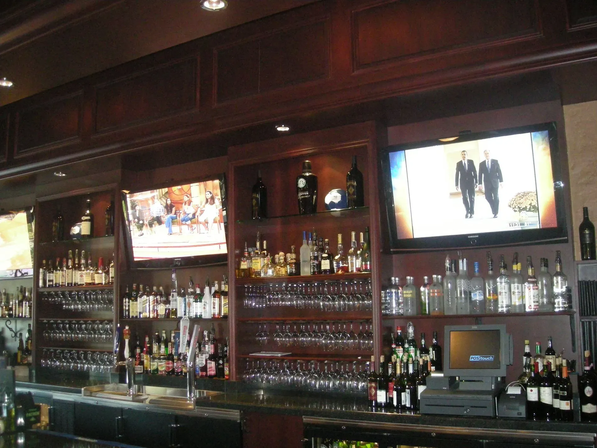 Dimly lit bar with shelves of liquor bottles, hanging glassware, and two TV screens above the counter.