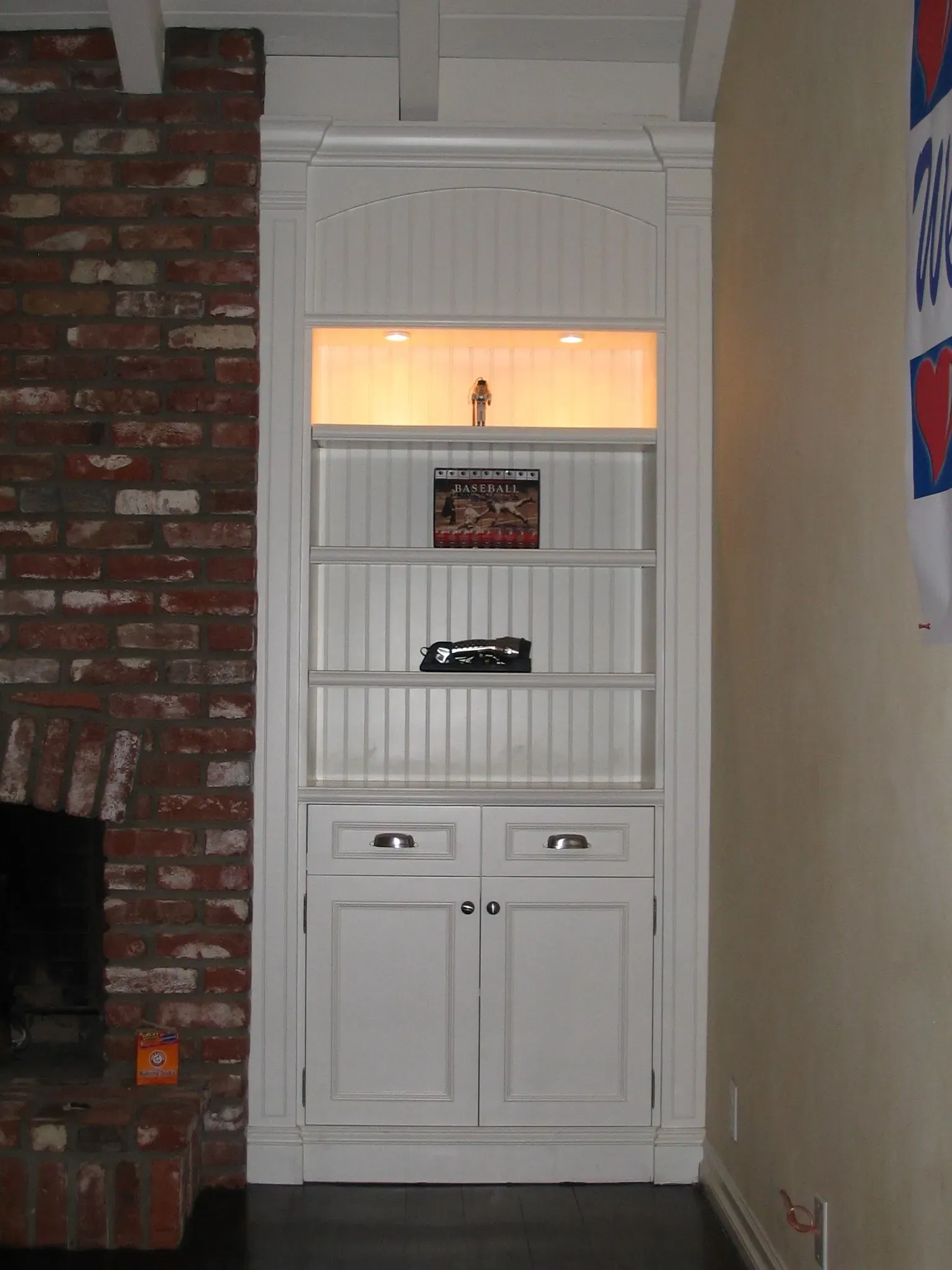 White built-in cabinet with lit glass shelf in a hallway, framed by brick and beige walls.