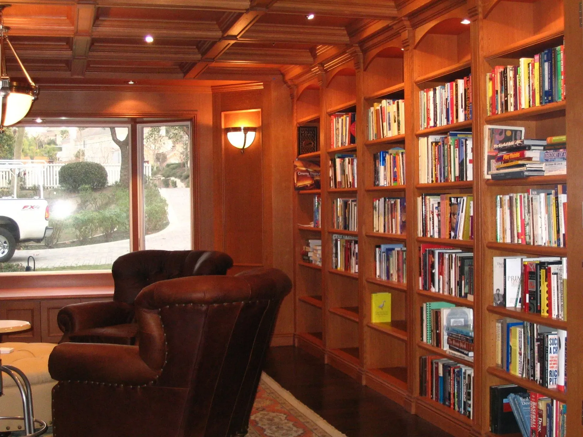 Warm wood-paneled library with bookshelves, leather chairs, and a window view outside.
