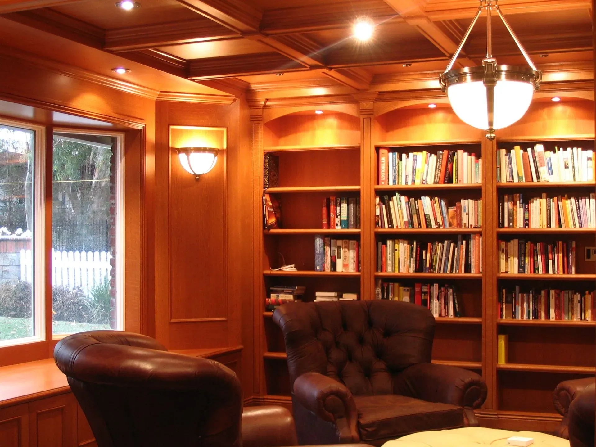 Warm wood-paneled library with bookshelves, armchairs, and a large window showing trees outside.