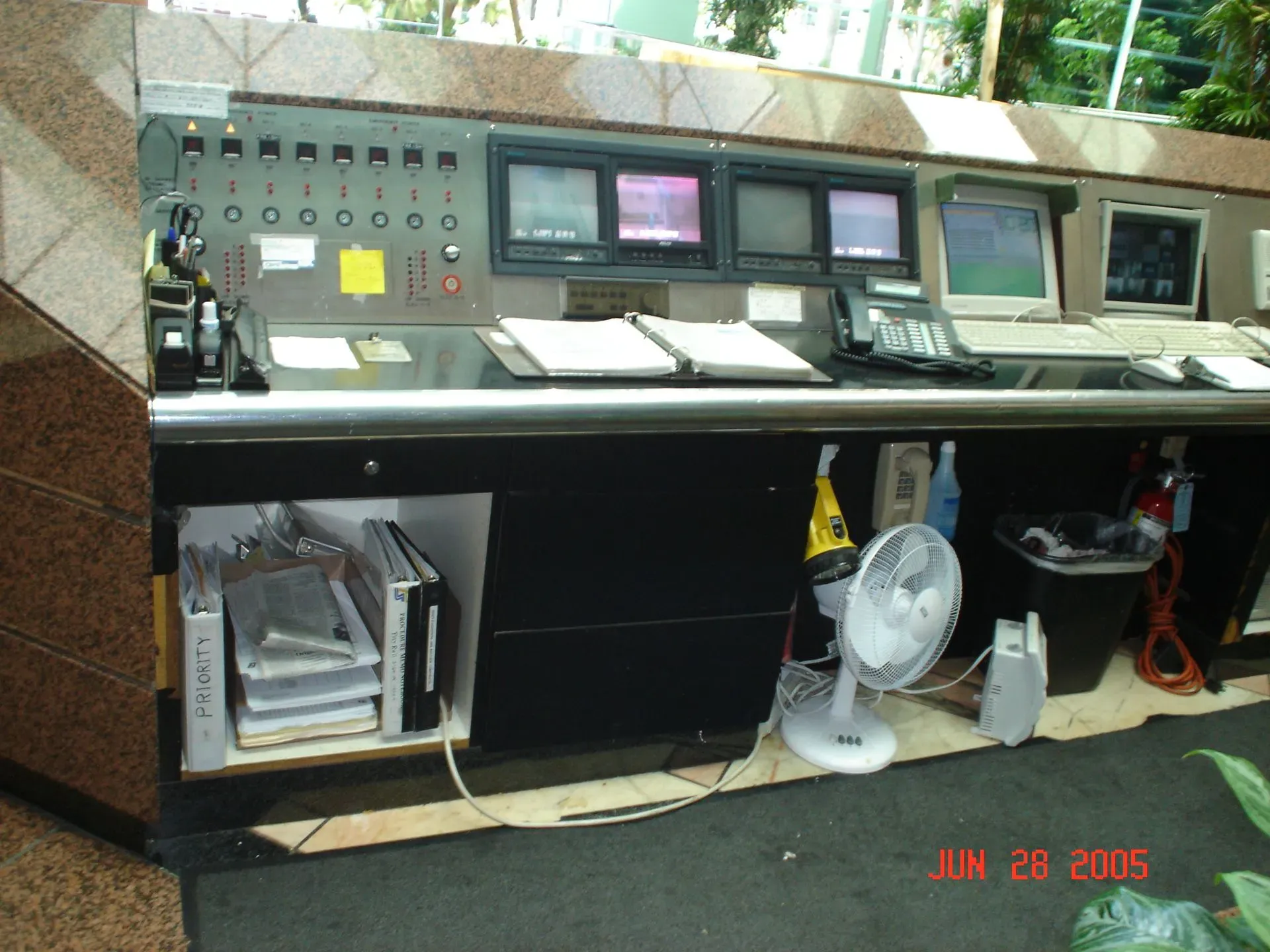 Indoor control room console with multiple monitors, buttons, papers, and a small fan beneath the desk.