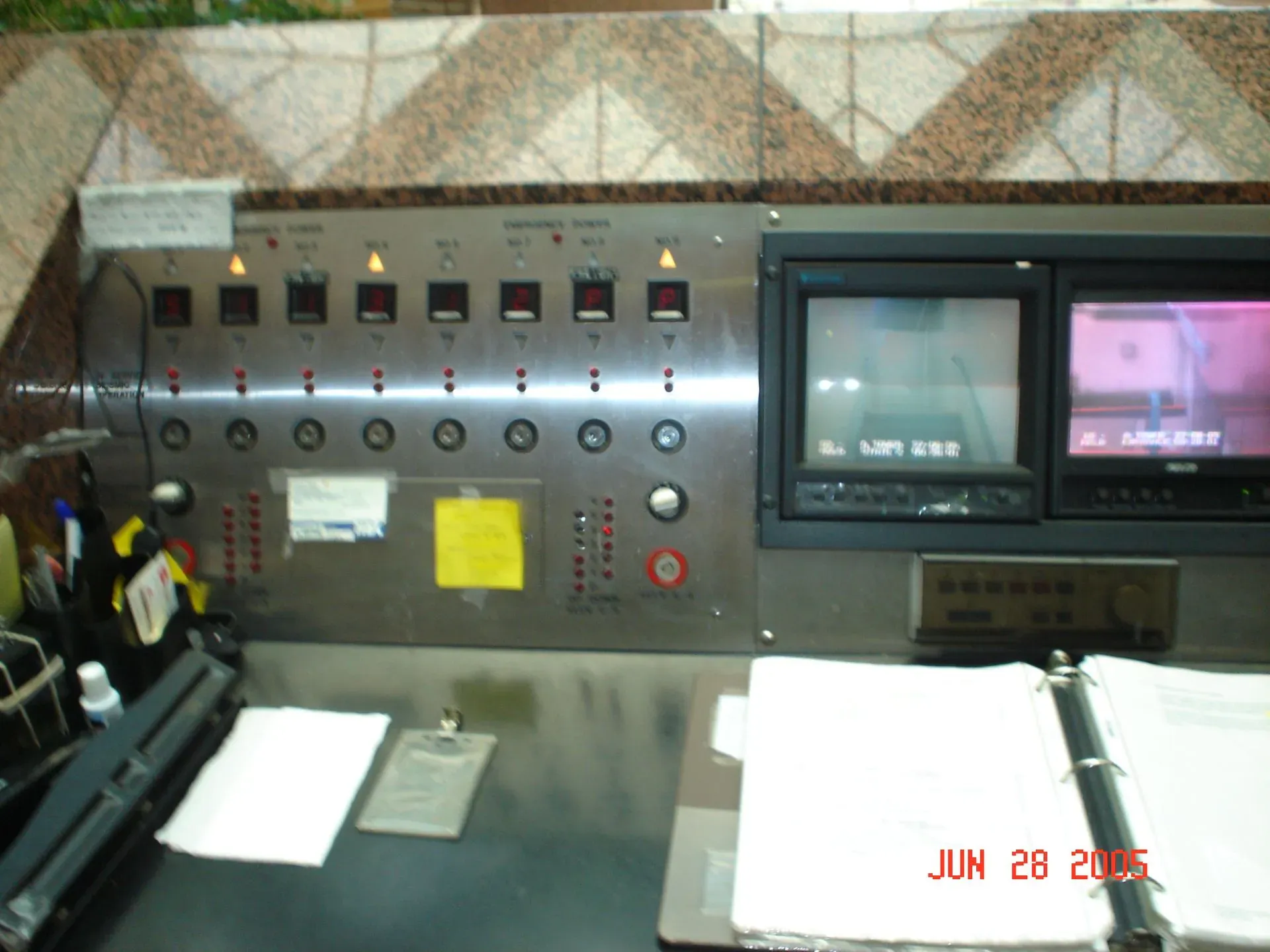 Control panel with switches and indicator lights beside two monitors in a dim industrial workstation.