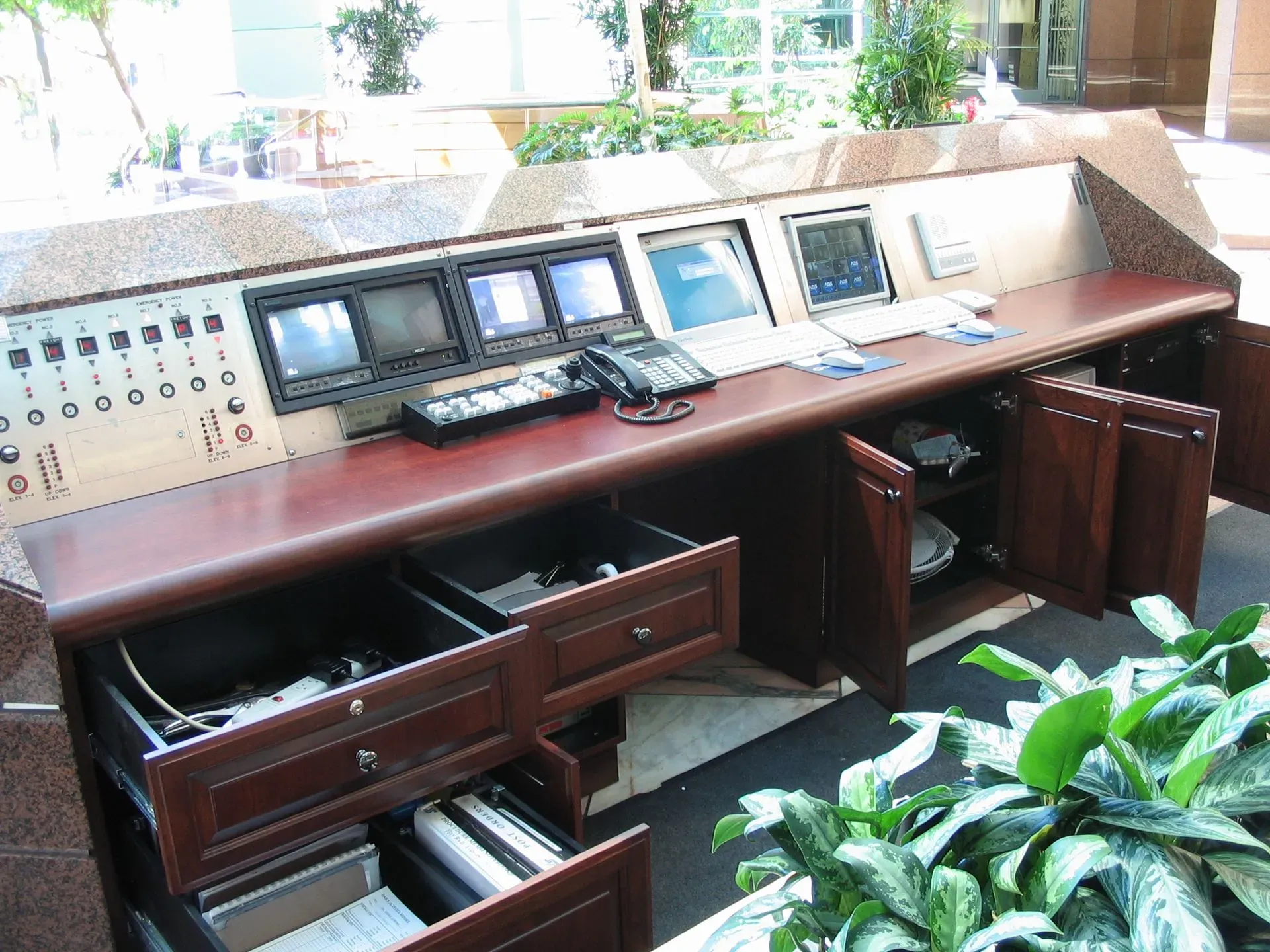 Wood-paneled control room with multiple monitors, keyboards, and open drawers beneath a long console.
