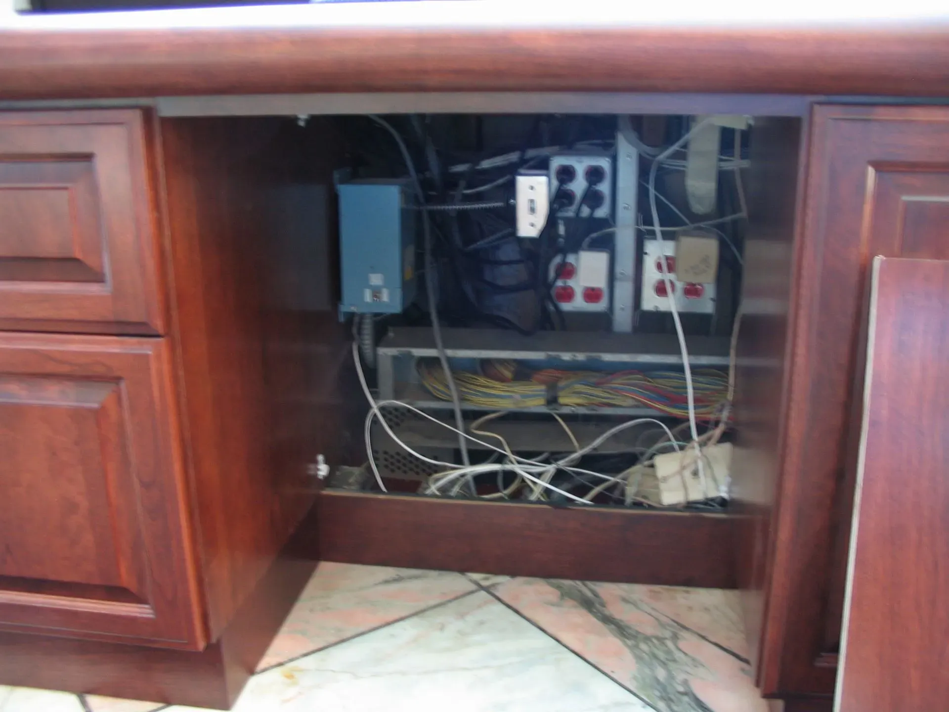 Open desk cabinet with exposed electrical wiring, outlets, and power strip inside.