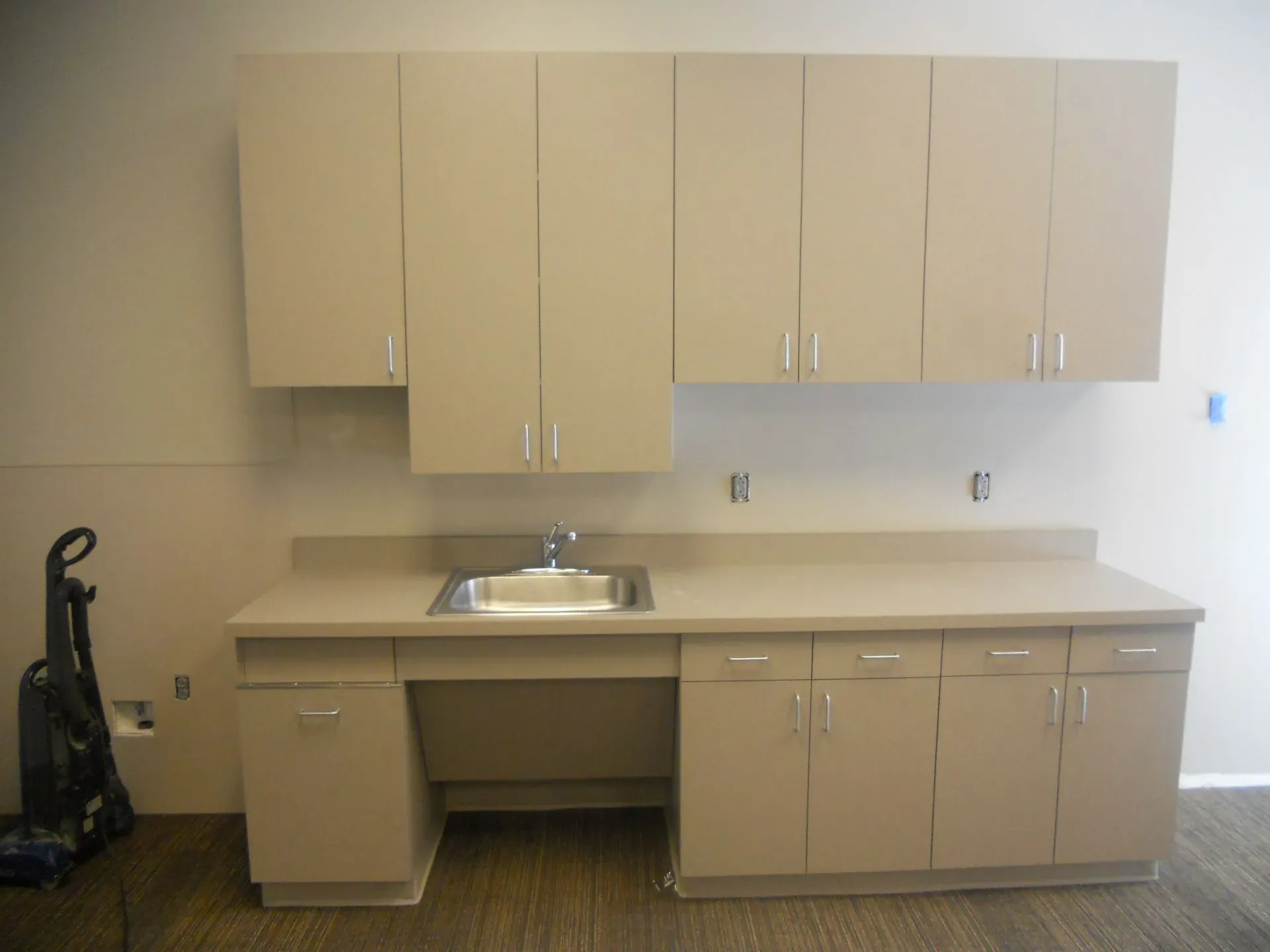 Empty break room with beige cabinets, countertop, and a small sink against a plain wall.